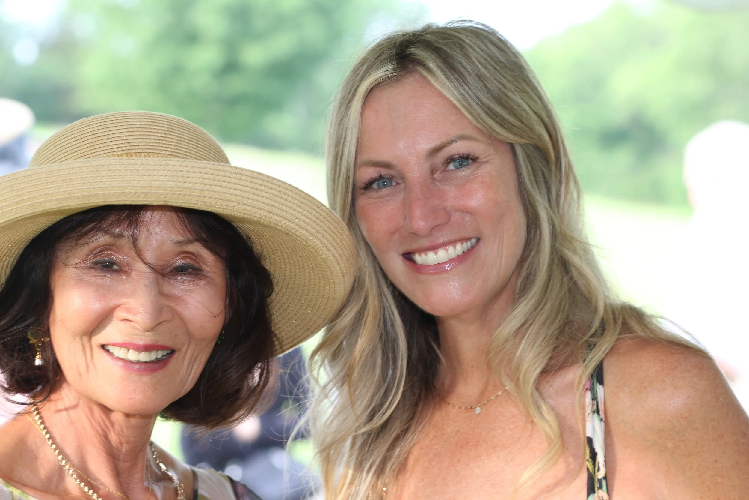 Two women smiling outdoors, one wearing a wide-brimmed straw hat and the other with long blonde hair, during daytime with blurred green background.