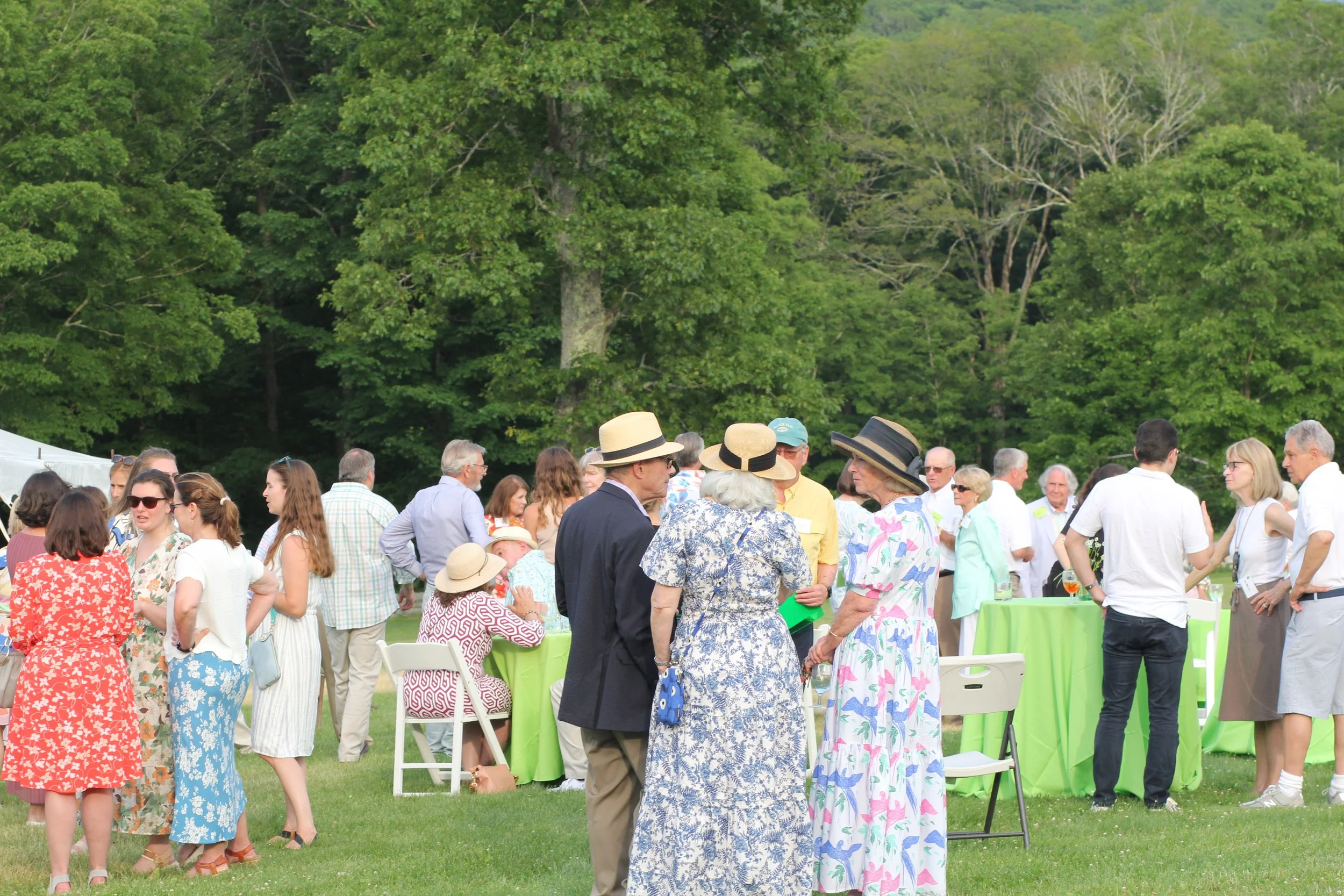 A group of people at an outdoor gathering or party, standing on green grass with many tables covered in green tablecloths. The attendees are dressed in casual and semi-formal summer clothing, with many wearing hats. The scene is set against a backdro