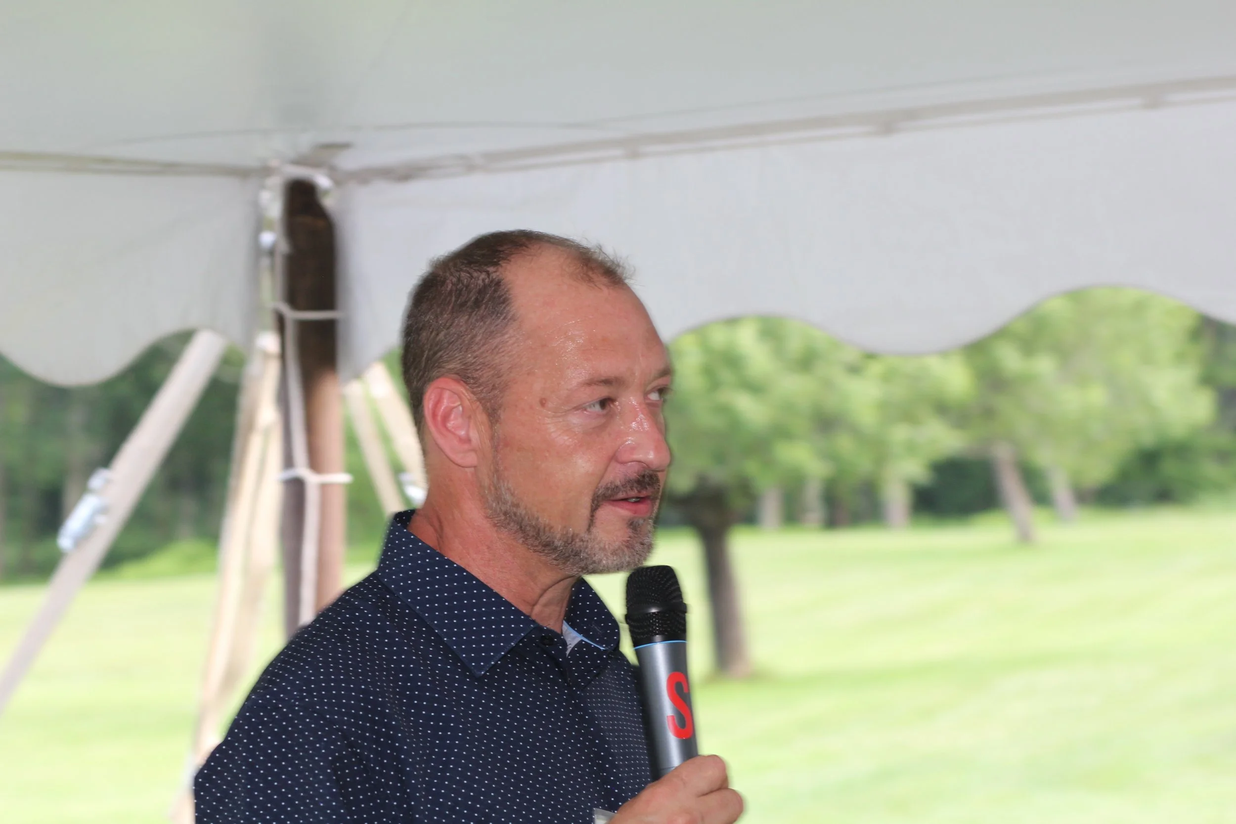 A man with short hair and a beard speaking into a black microphone during an outdoor event, with a green field and trees in the background.