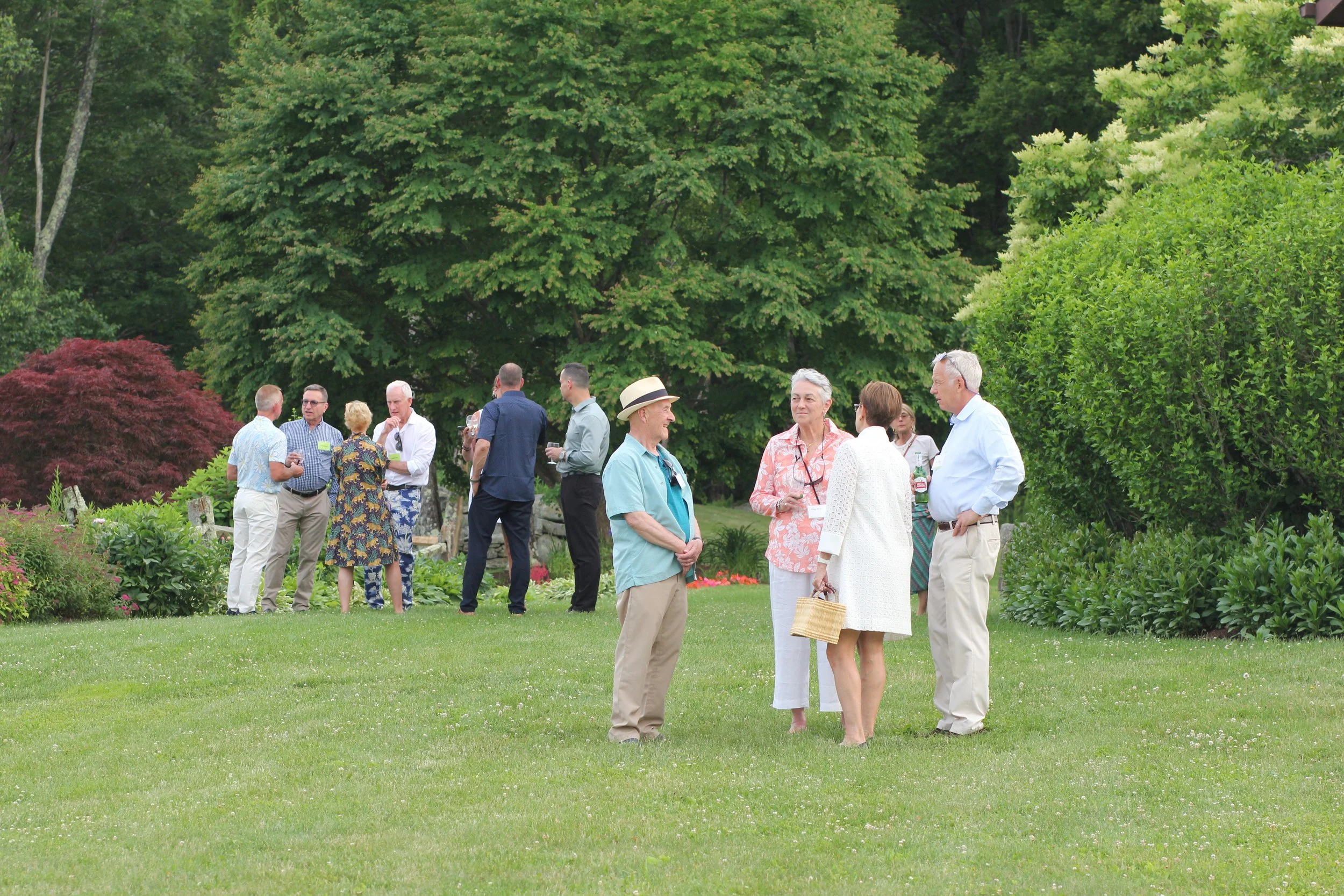 People are gathered outdoors in a lush green garden, chatting and socializing during what appears to be a casual outdoor gathering or event.