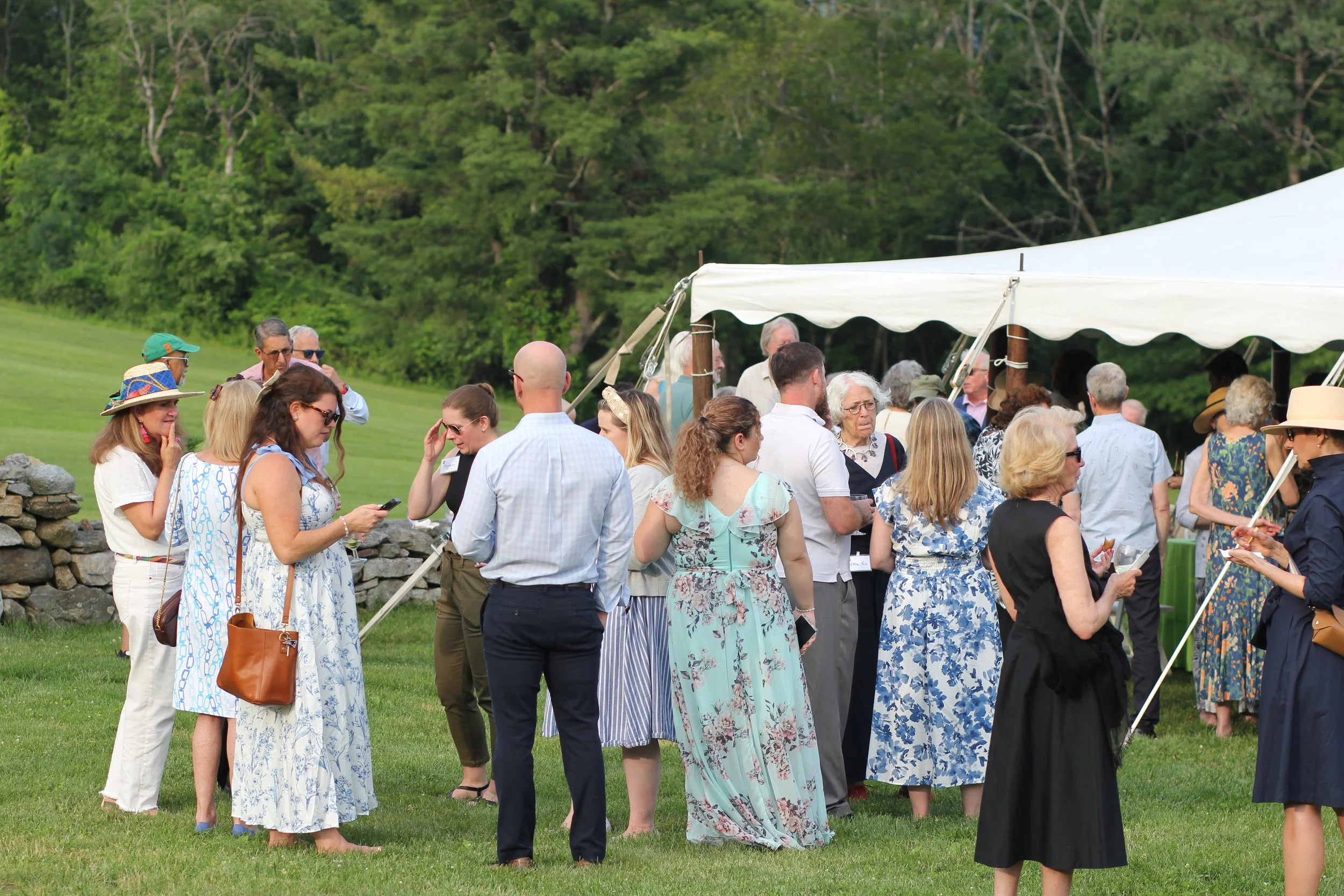 A group of people gathered outdoors near a large white tent on a grassy field, with trees in the background. Some individuals are dressed in casual and summer clothing, and some are wearing sunglasses or hats.