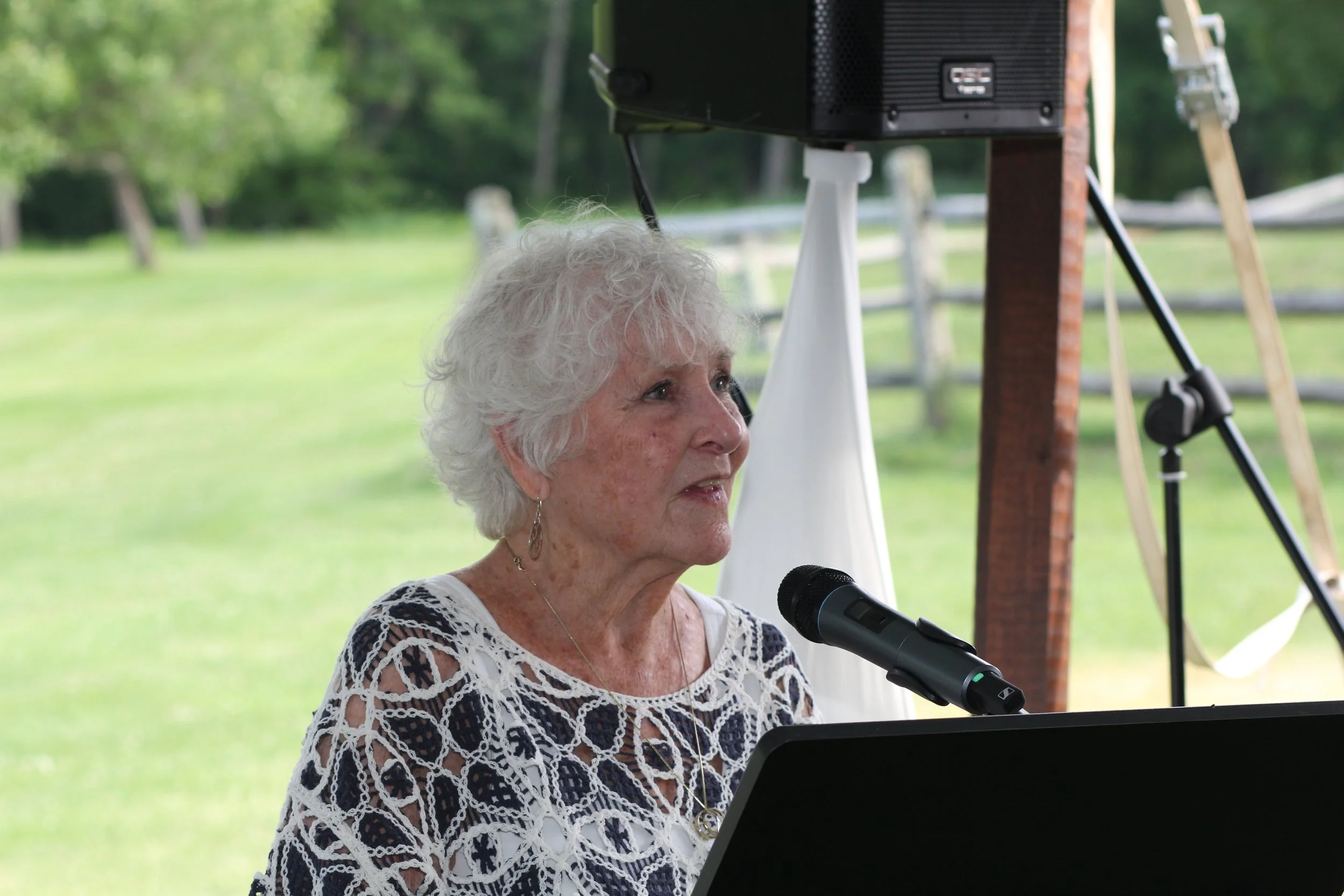 An elderly woman with curly white hair speaking into a microphone while playing a piano outdoors, with green trees and a wooden fence in the background.