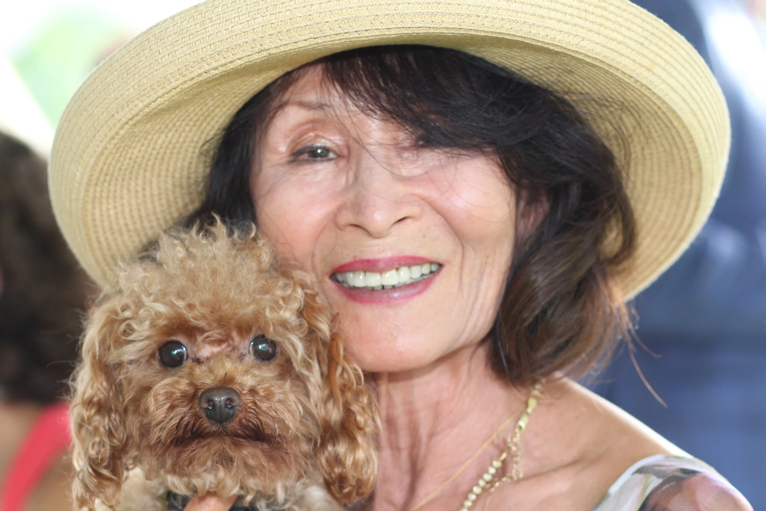 An elderly woman in a straw hat smiling while holding a small, curly-haired brown puppy close to her face.