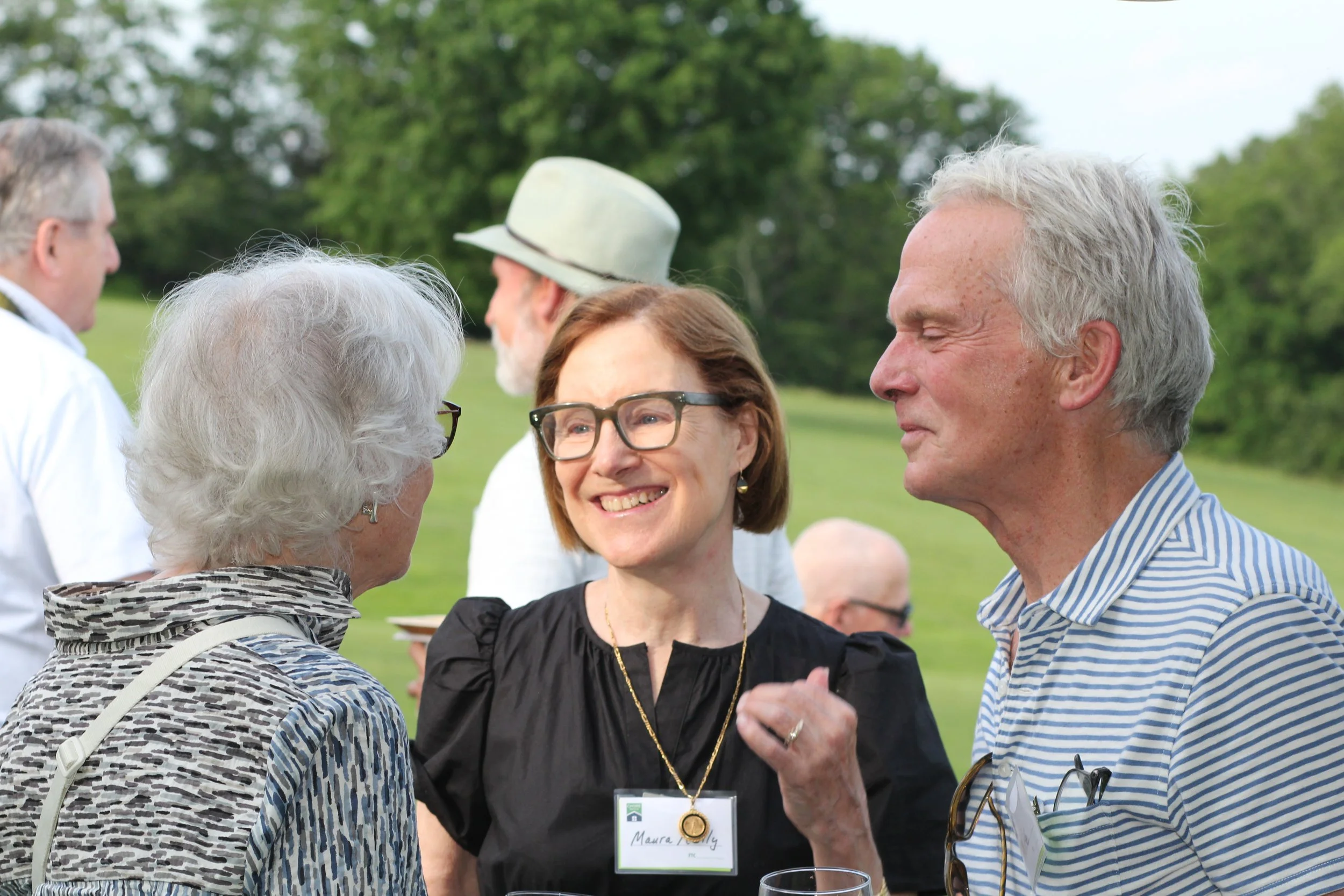 A group of five older adults outdoor at a social event, engaged in conversation, with a green lawn and trees in the background.