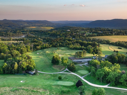 Aerial view of a lush golf course surrounded by trees, with rolling hills in the background during sunset.