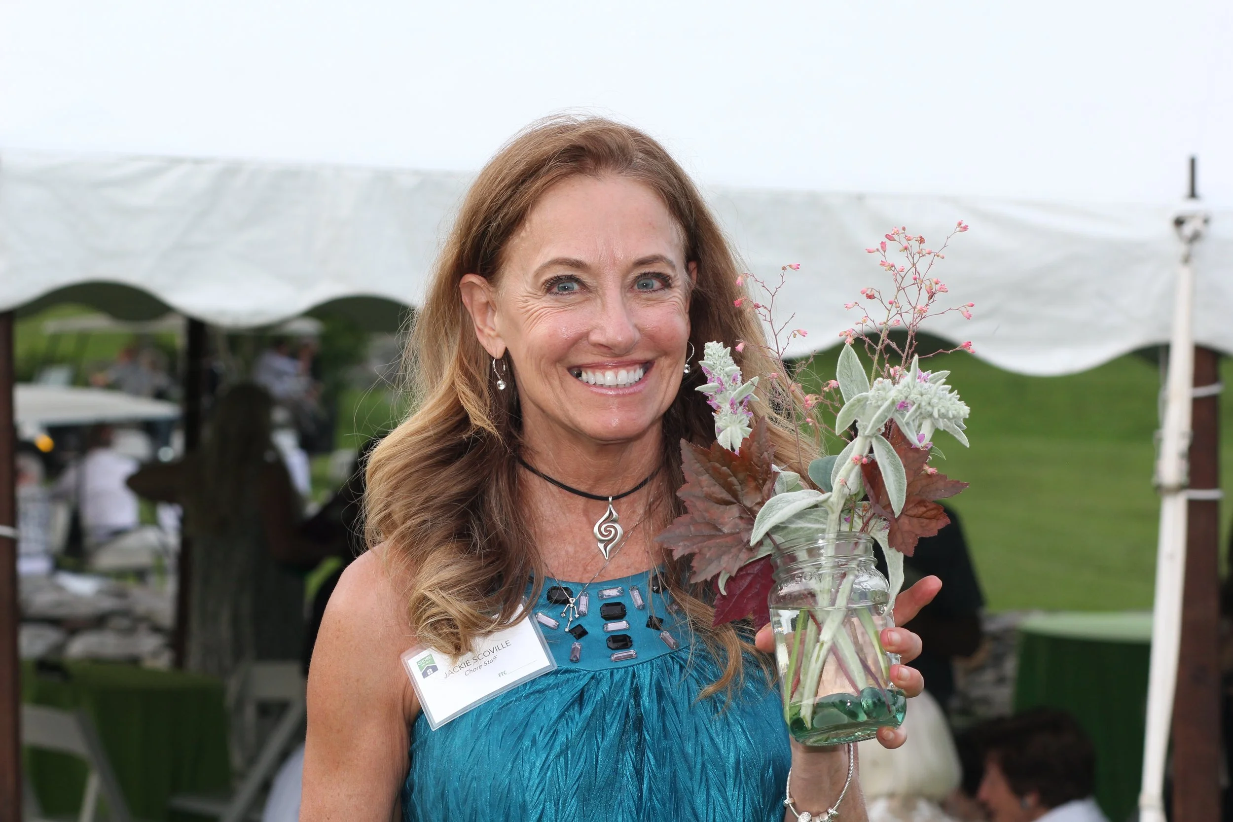 A smiling woman with long brown hair holding a glass jar with a bouquet of flowers, wearing a blue dress and a choker necklace, at an outdoor event in Litchfield County with a white tent in the background.