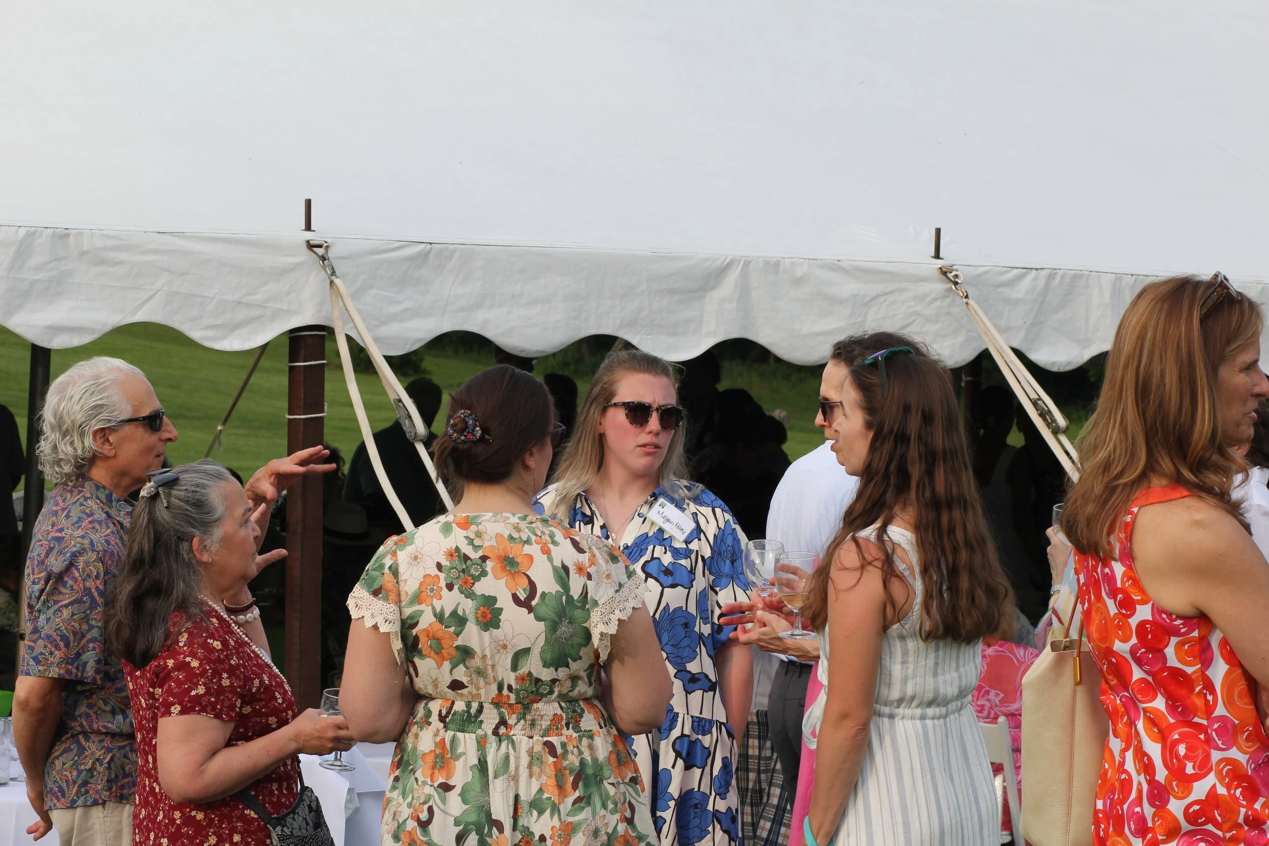 Group of people at an outdoor fundraiser in Litchfield County for Chore Service under a white canopy, engaging in conversation, holding drinks, wearing casual summer clothing.