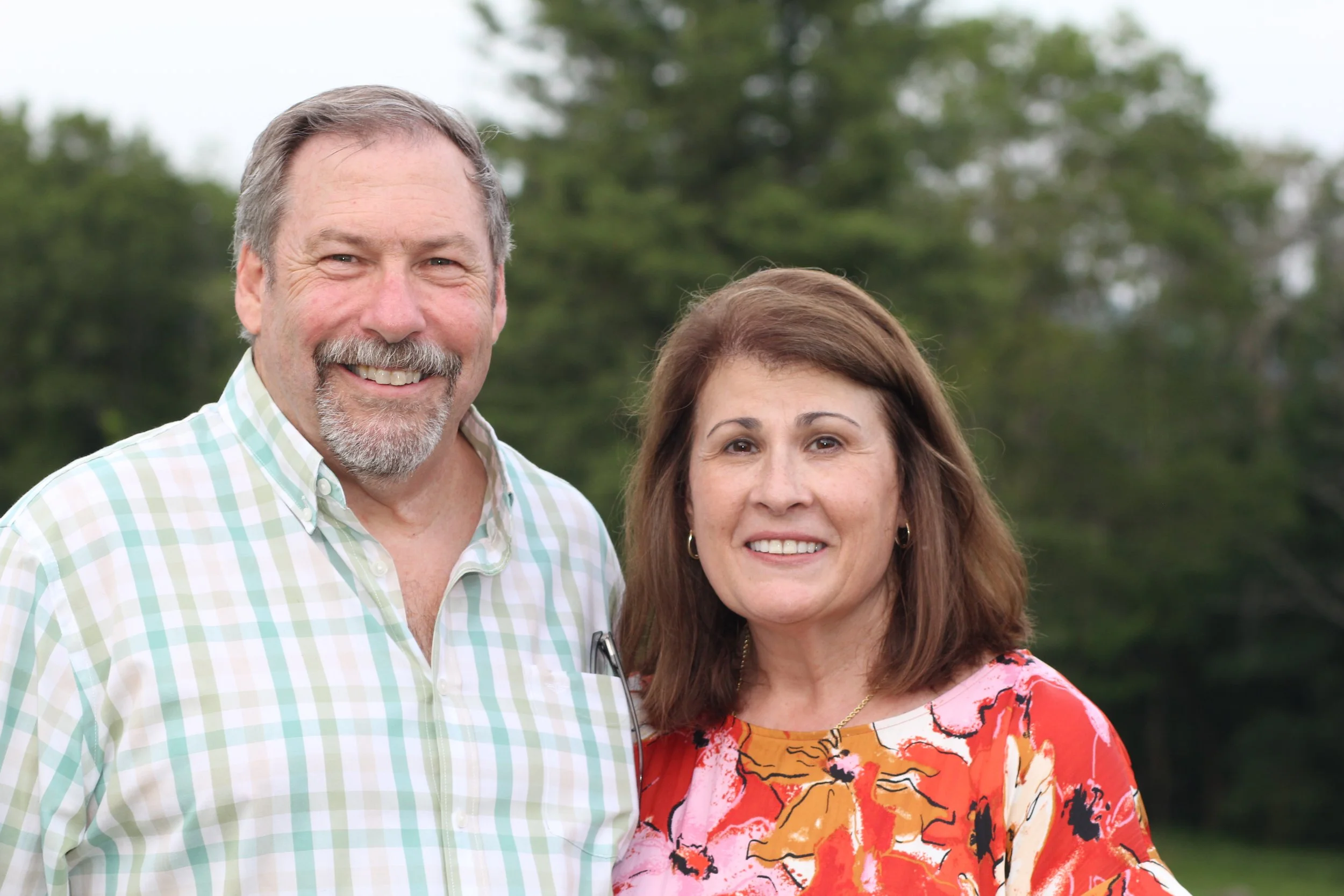 A smiling middle-aged man with gray hair standing next to Chore Service's Executive Director, Jane Maclaren, a smiling middle-aged woman with shoulder-length brown hair wearing a colorful floral dress, outdoors with trees in the background.