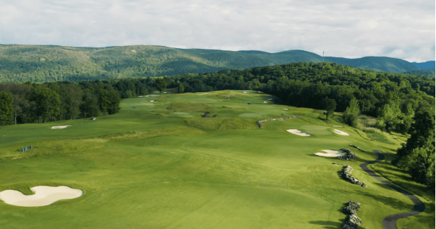 Aerial view of a lush green golf course with sand traps and a winding path, surrounded by trees and distant mountains under a cloudy sky.