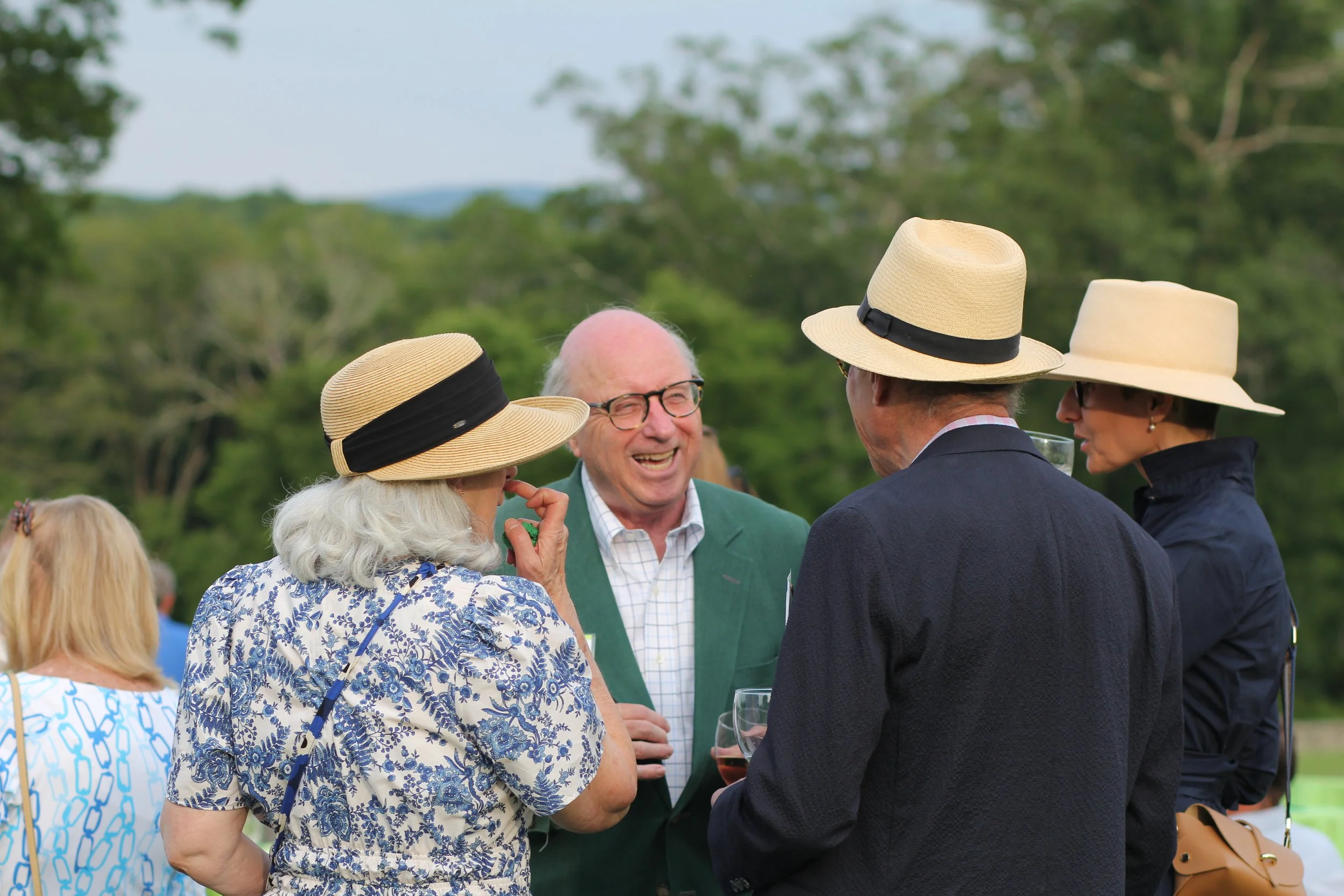 A group of four elderly people, two women and two men, dressed in semi-formal summer attire, engaged in conversation outdoors, with trees and a scenic landscape in the background, at a fundraiser in Litchfield County for Chore Service .