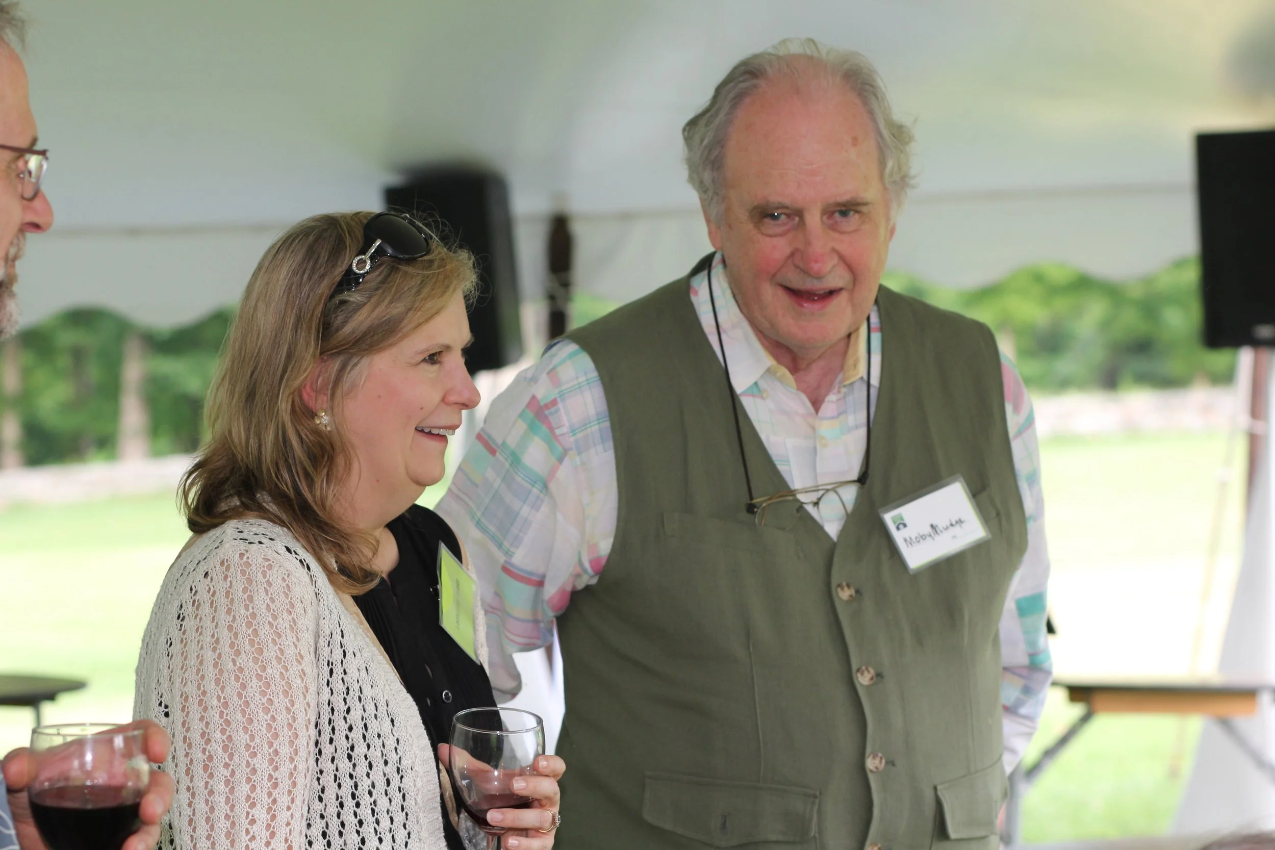 A group of people laughing and talking at an outdoor event in Litchfield County, with wine glasses in hand, under a tent with a scenic green landscape in the background.