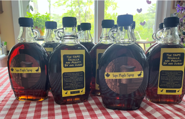 Six bottles of homemade maple syrup on a red checkered tablecloth in front of a window with green trees outside.