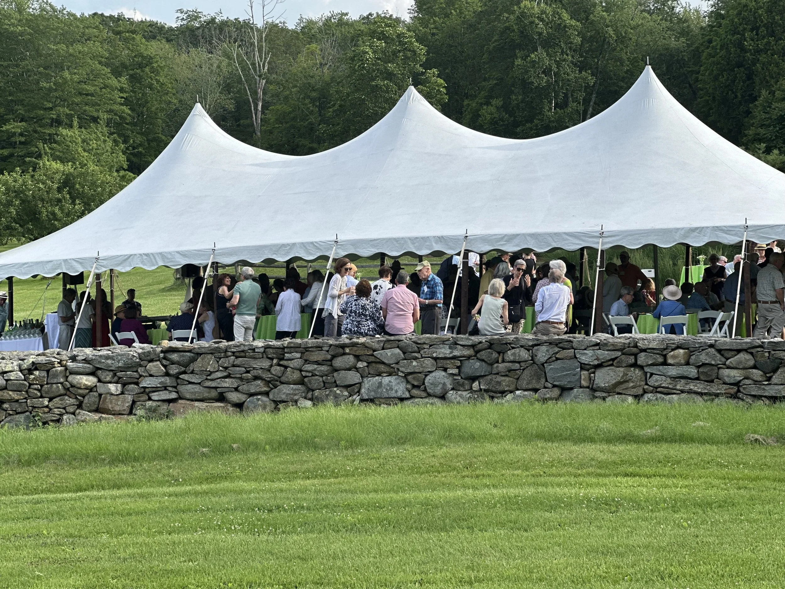 Outdoor event under a large white tent with people gathered, alongside a stone wall and green grassy field, in a lush, wooded area.