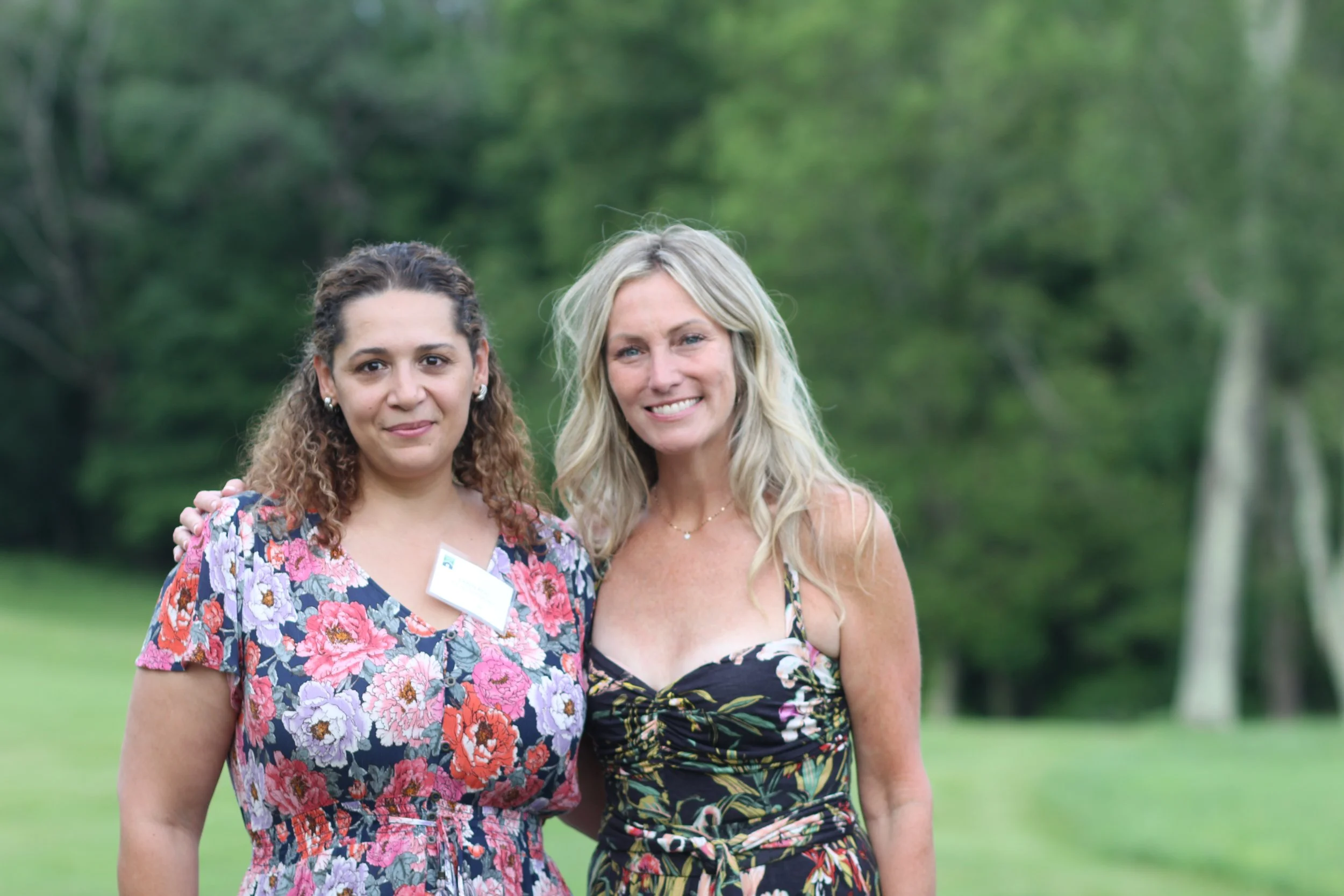 Two women standing outdoors in front of a lush green background in Litchfield County, smiling at the camera. One woman has curly dark hair wearing a floral dress with a name tag. The other woman has long blonde hair is wearing a black floral dress.
