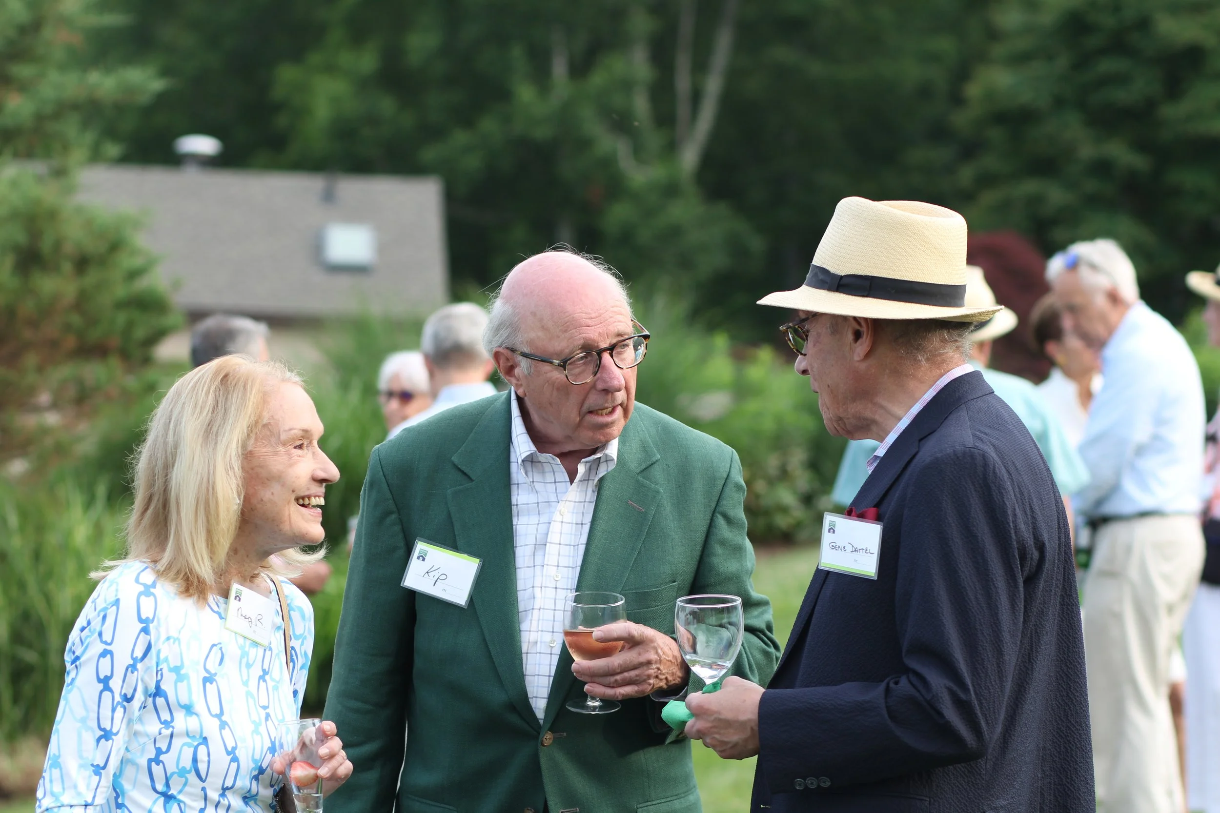 Three seniors enjoying a conversation at an outdoor gathering, with two men and a woman smiling and holding wine glasses, in a lush green garden setting.