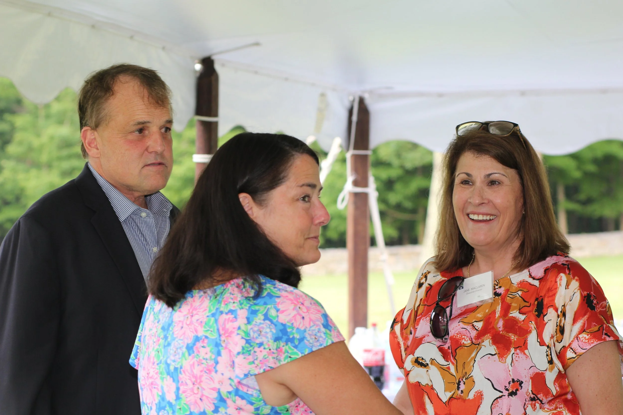 Three adults engaged in conversation at an outdoor event in Litchfield County for Chore Service under a tent, with a background of trees and greenery.