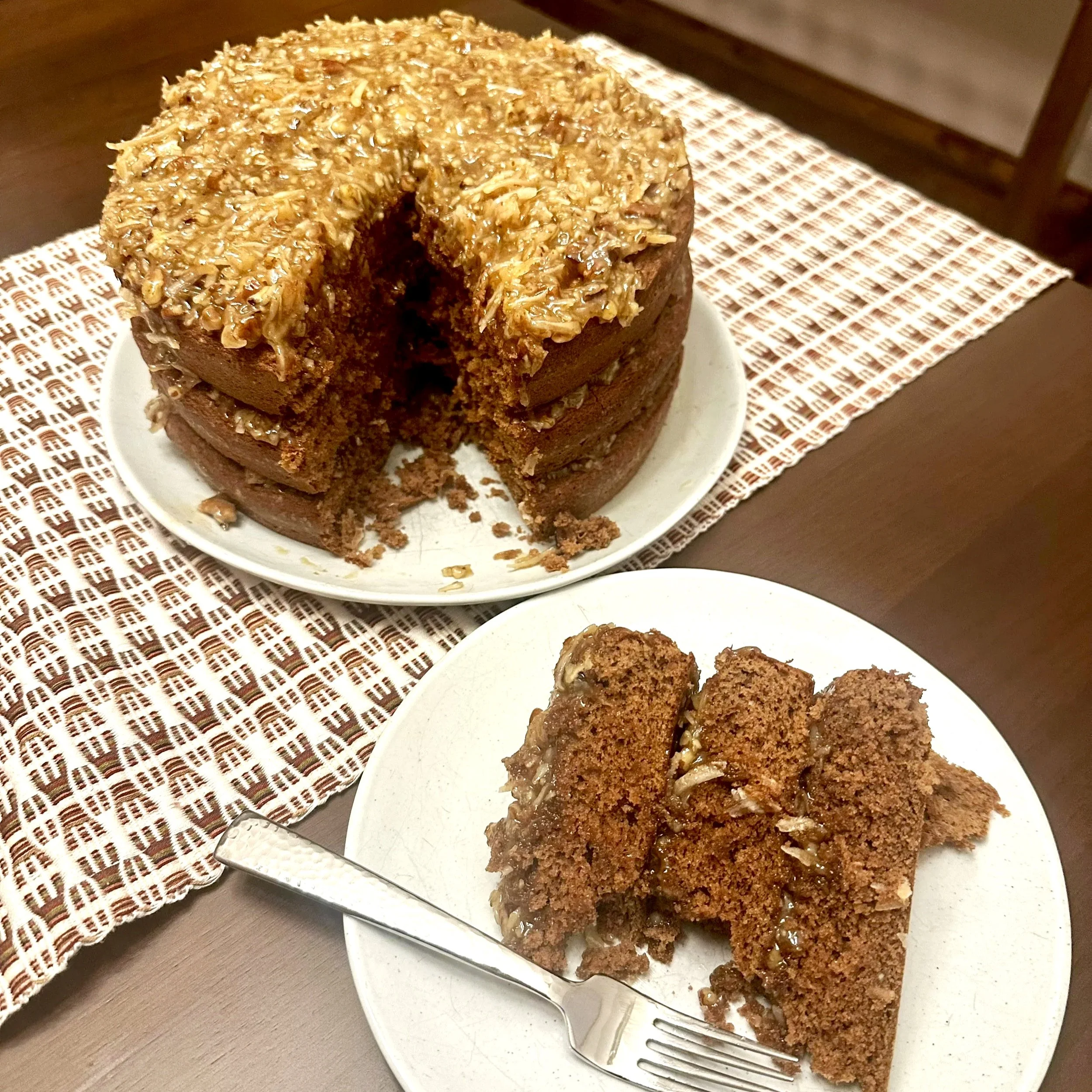 Three-tiered cake with a slice removed, revealing soft layered interior, set on a plate beside a silver fork, with crumbs and frosting details visible.