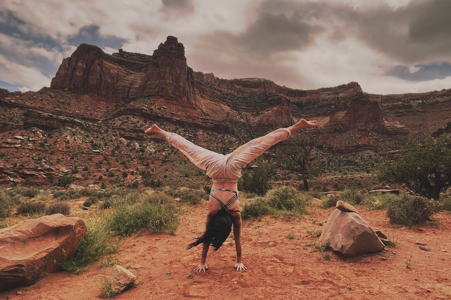 A woman in a joyful handstand on red desert sand, mountains and dramatic sky in the background
