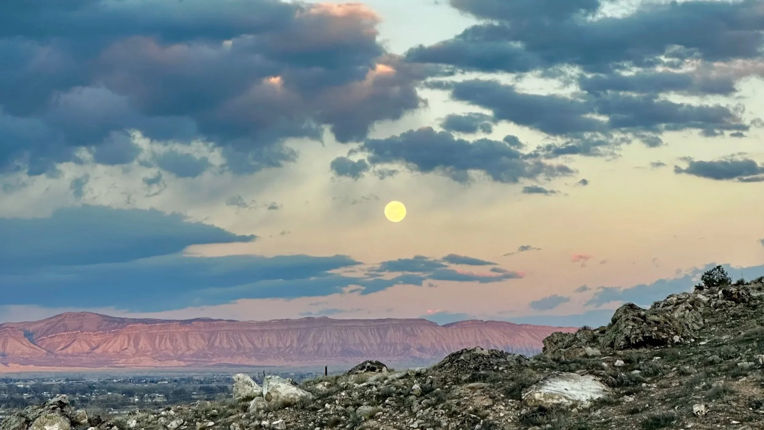 Full moon rising above a mountain ridge at sunset, with soft blue and pink hues blending across the evening sky and the dark silhouette of the peaks beneath the glowing orb.