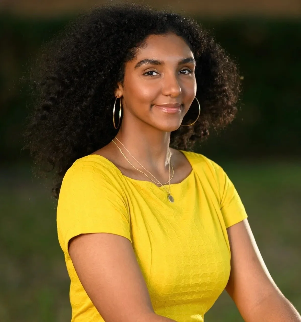 Young woman with curly hair wearing a yellow dress and jewelry, smiling outdoors.