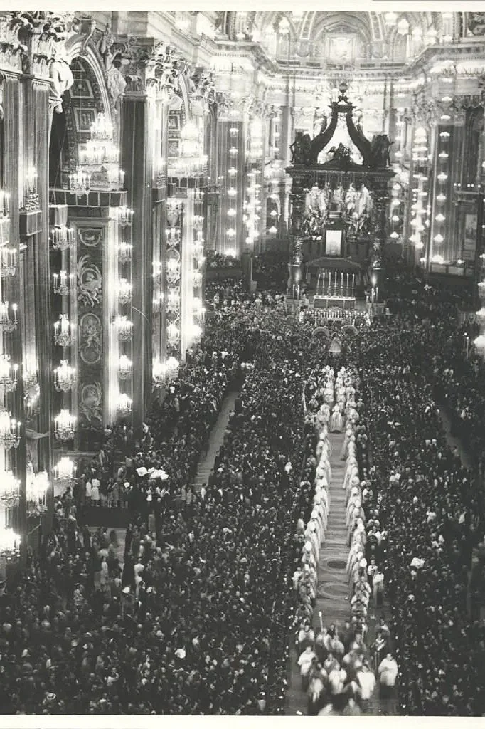 A large religious gathering inside an ornate church or cathedral with a procession of people in white robes and a large congregation, with high ceilings, chandeliers, and sculptures.