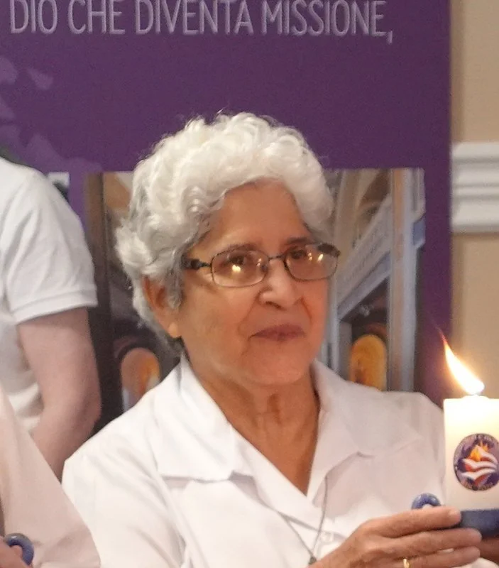 An elderly woman with short, curly white hair and glasses holding a lit candle. She is wearing a white shirt, standing in front of a purple backdrop with Italian text.