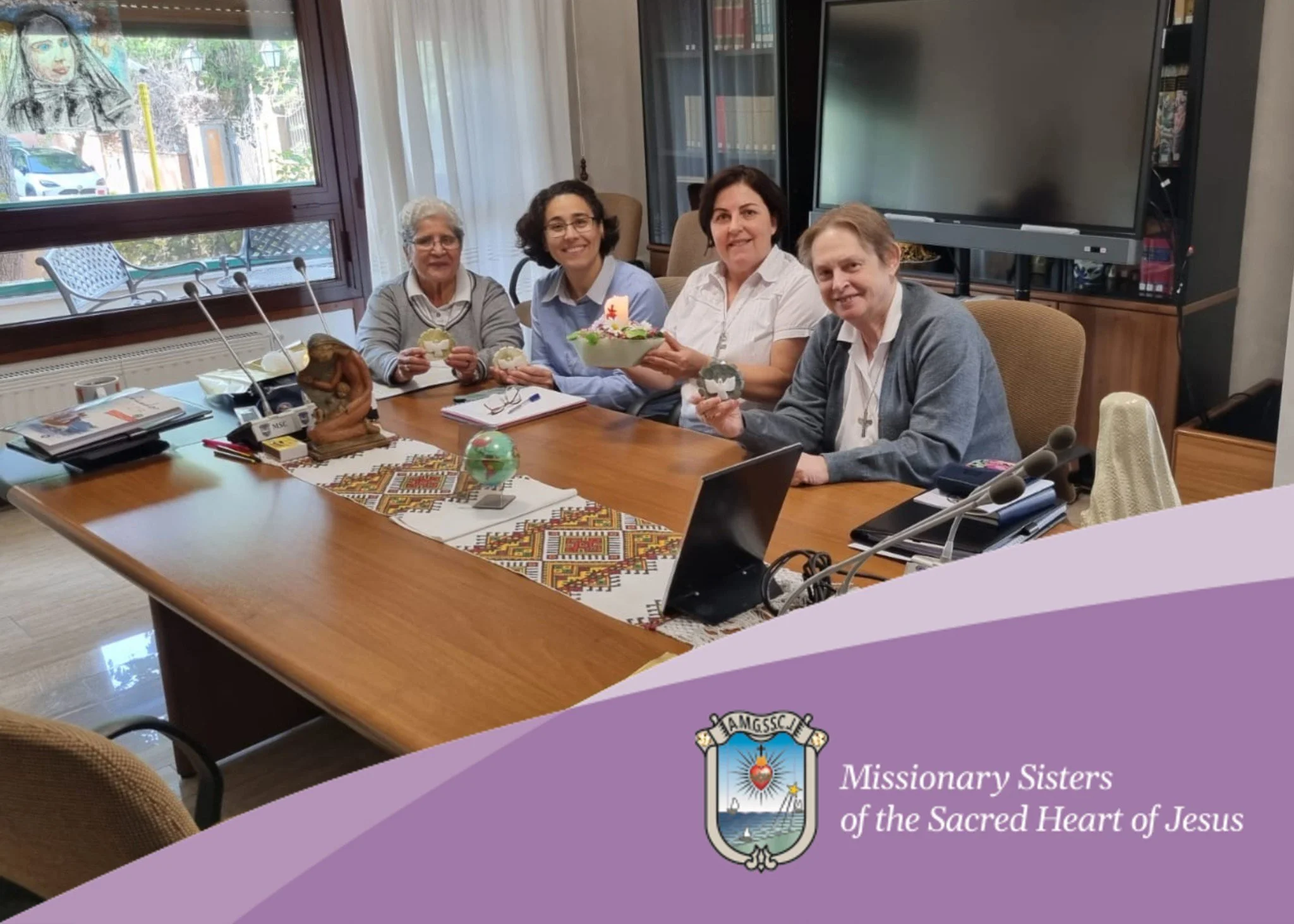 A group of five women sitting around a large conference table in a room, smiling and looking at the camera. The table has documents, a colorful embroidered cloth, and some decorative items, including a small globe. Behind them are windows with curtains and a large TV screen. The image has a purple overlay with a logo and text that reads 'Missionary Sisters of the Sacred Heart of Jesus'.