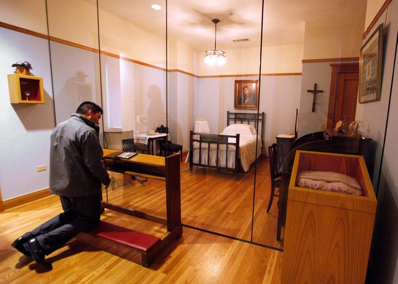 A man in a gray jacket is kneeling down with one knee on the floor and one foot on the ground, appearing to be pushing a wooden framed glass-encased bench in an interior room. The room has a bed, a painting, a crucifix on the wall, and various furniture and decorations.