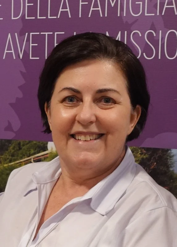Smiling woman with short dark hair wearing a white shirt, standing in front of a purple background with partial text.