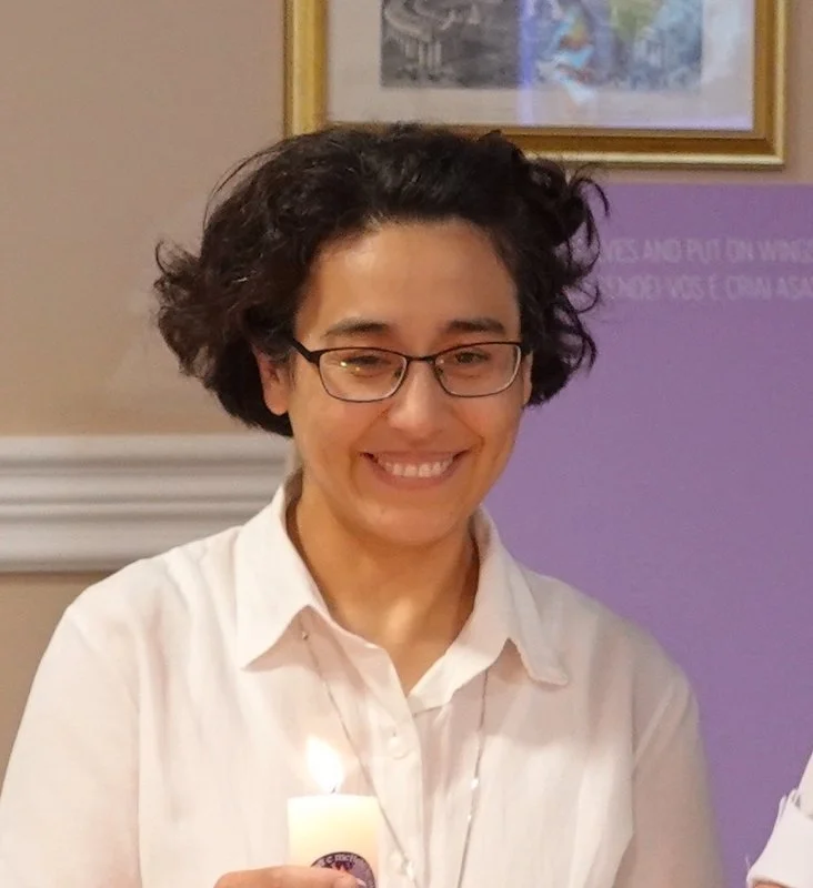A woman with short curly dark hair, glasses, and a white shirt smiling, holding a lit candle.