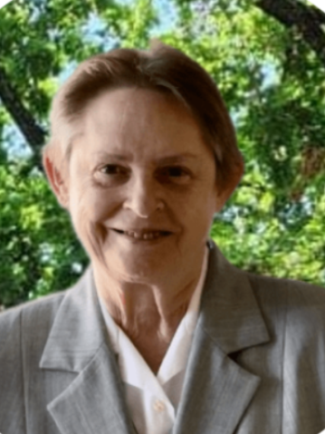 An elderly woman smiling outdoors with green trees and foliage in the background, wearing a gray blazer and white shirt.