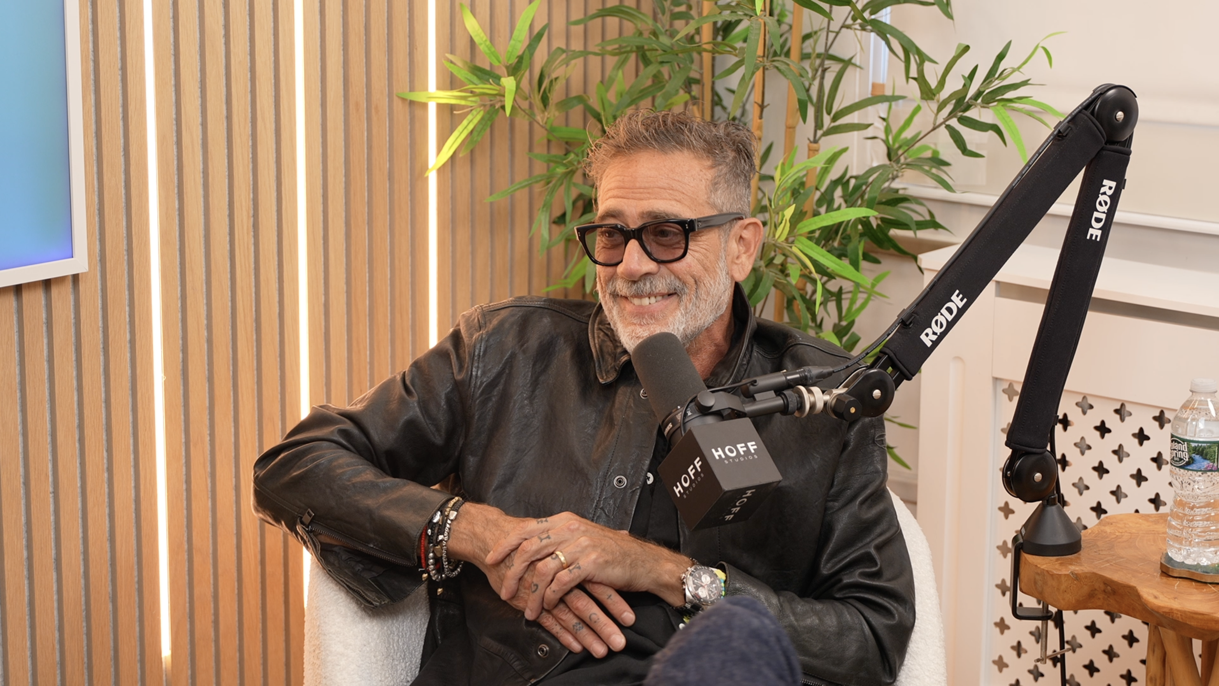 A middle-aged man with gray hair and beard, wearing black glasses and a black leather jacket, smiling during a recording session with a Rode microphone in front of him. There are green plants and wooden slats in the background.