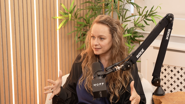 A woman with long, curly blonde hair sitting in a recording studio, speaking into a microphone with a Rode arm, surrounded by green plants and wooden paneling.
