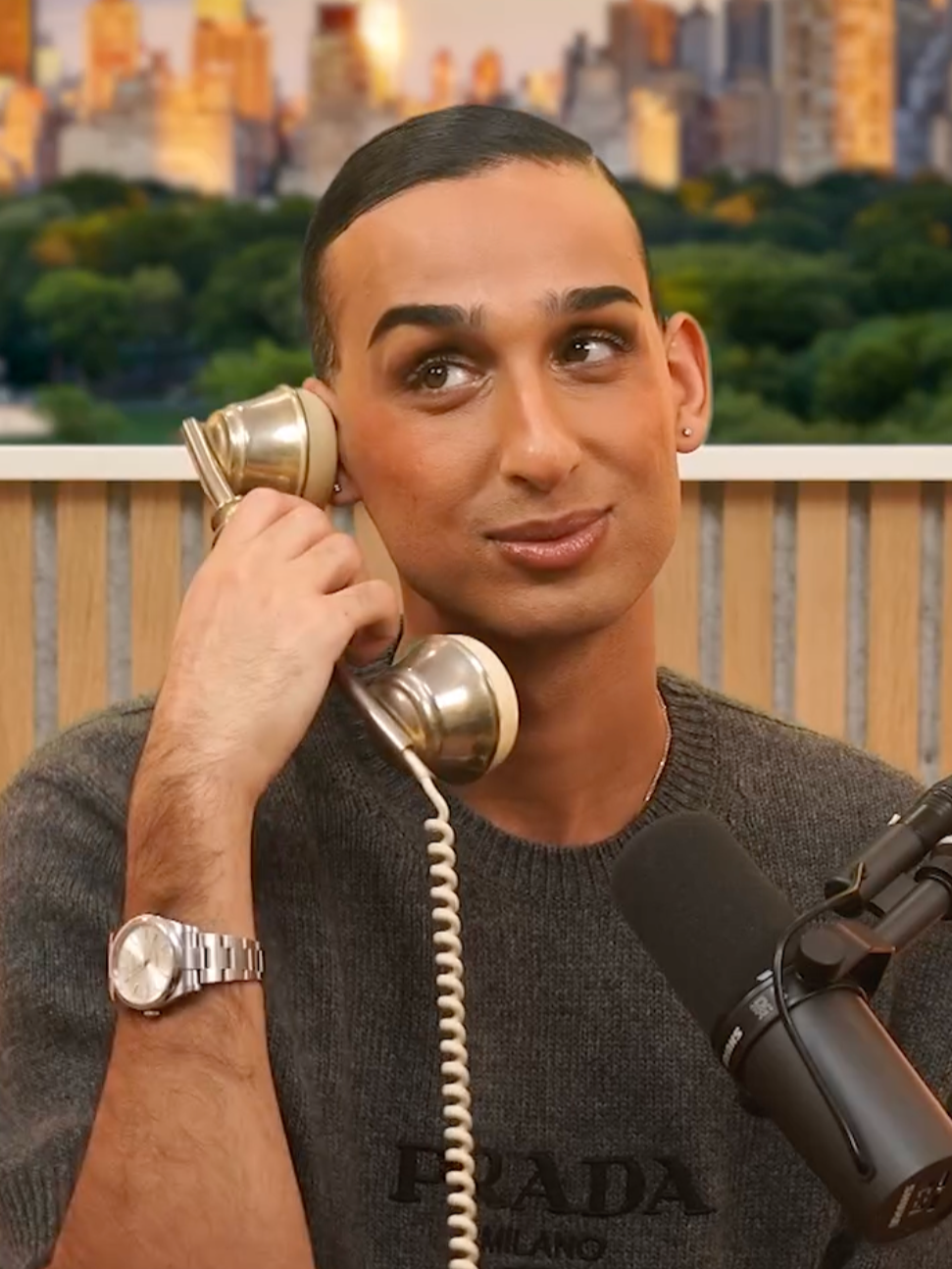 A young man with dark hair and eyebrows, wearing a Prada sweatshirt and a silver wristwatch, holds a vintage rotary phone to his ear, sitting in front of a microphone with a blurred cityscape background.