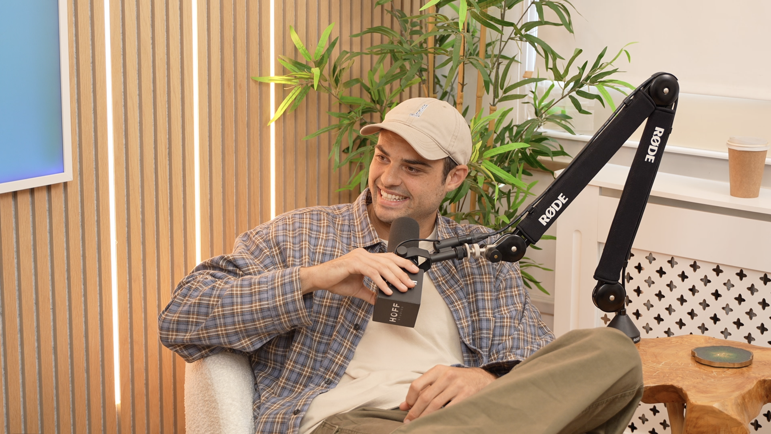 A man wearing a beige cap and plaid shirt smiling, speaking into a microphone, in a room with wooden vertical panel walls and green plants.