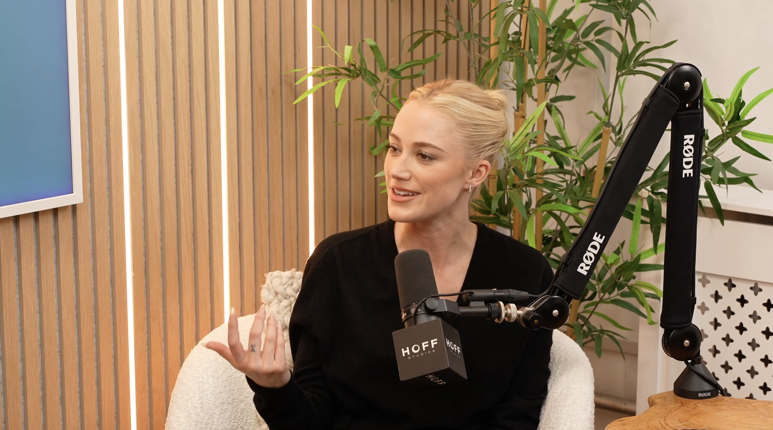 A woman with blonde hair in a bun, wearing a black sweater, speaking into a professional microphone labeled 'HOFF' in a recording studio or podcast setting. She is gesturing with her left hand. Behind her, there are green plants and a wooden wall with vertical slats.