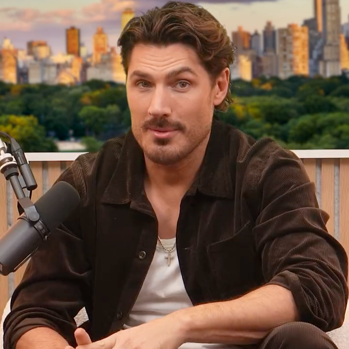 A man with dark hair and a beard, wearing a brown jacket and a white shirt, sitting at a table with a microphone, with a city skyline and green park in the background.