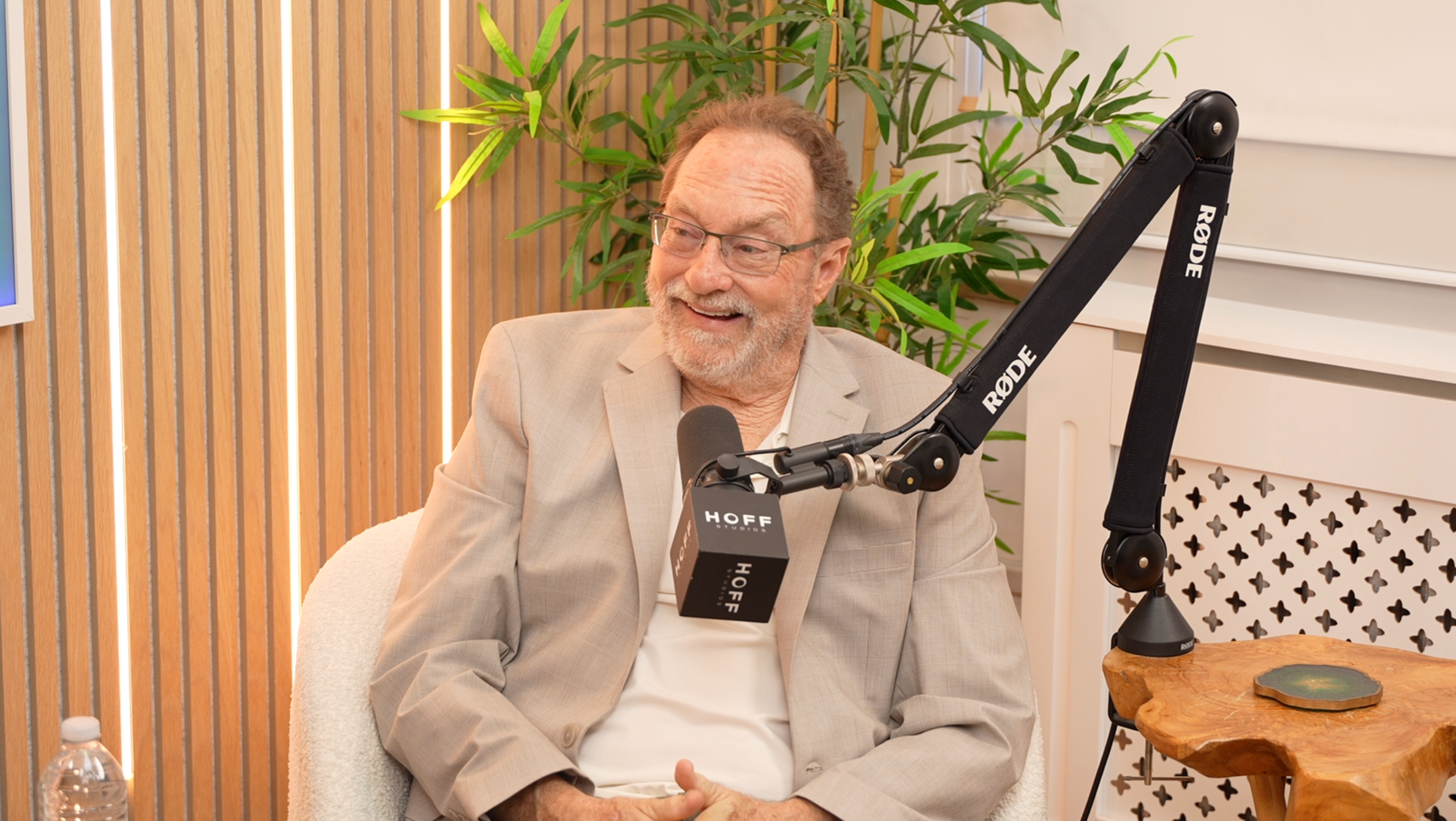 An older man in glasses and a beige blazer sitting at a table, smiling during a recording interview with a RODE microphone in a studio with wooden wall paneling and green plants in the background.