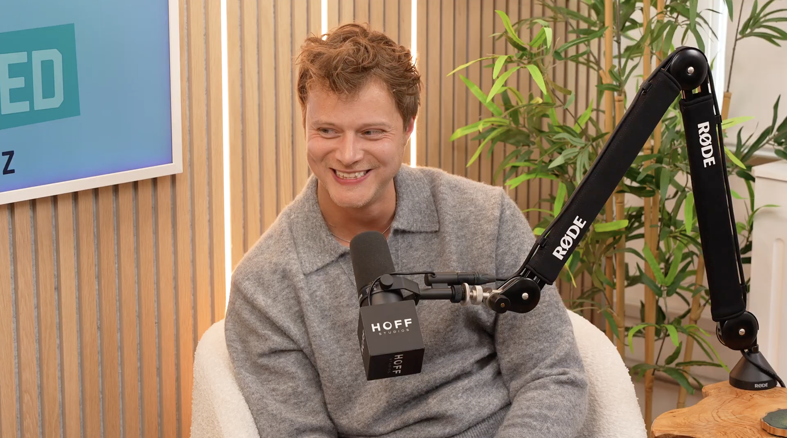 A man with short, wavy hair smiling during a podcast recording, seated in front of a microphone labeled 'HOFF Studios' with a 'RODE' microphone arm, in a room with a wooden wall, green plants, and a blue screen.