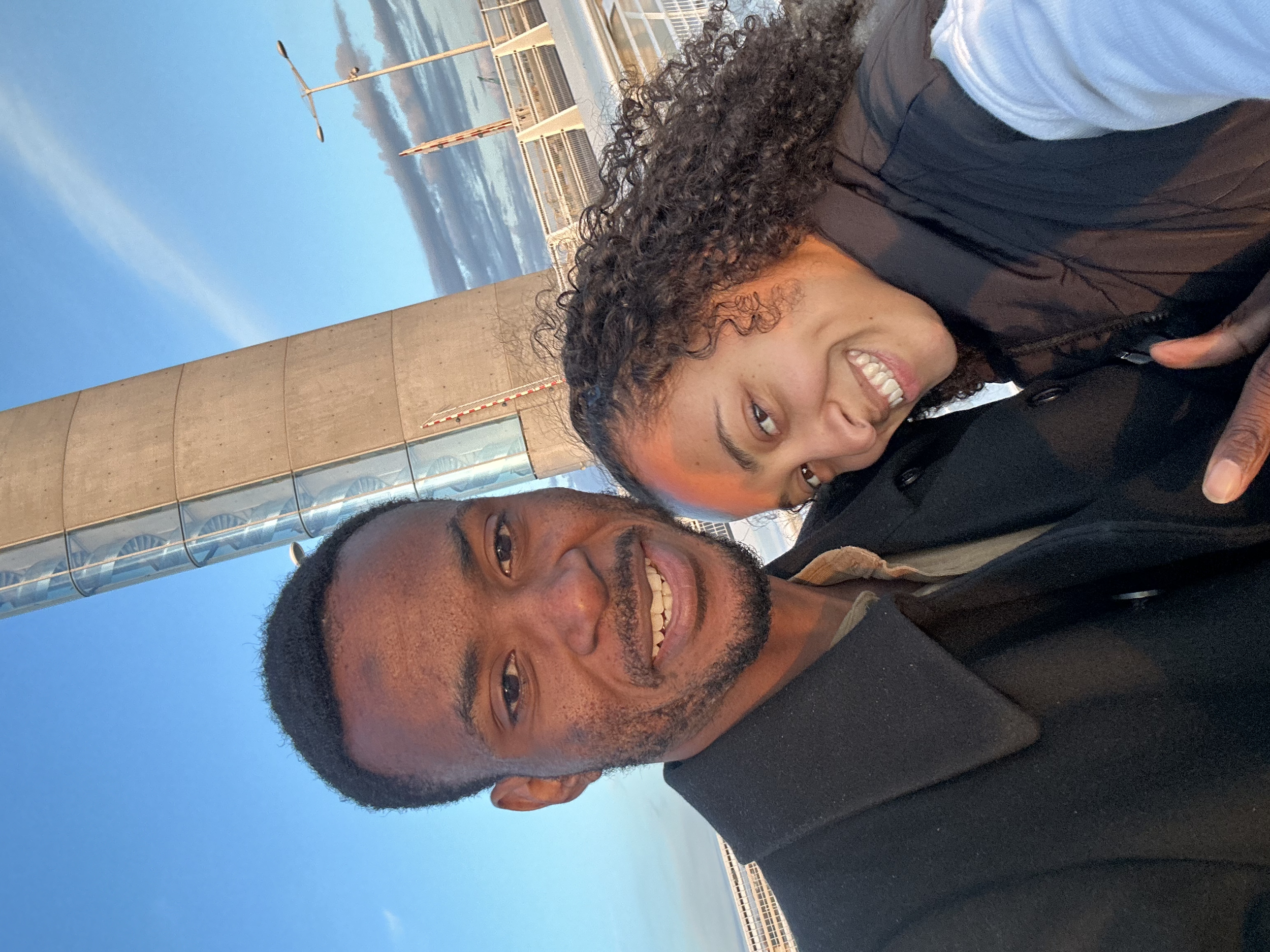 Deux personnes souriantes prennent un selfie devant une structure en béton et en verre, avec un ciel bleu en arrière-plan.