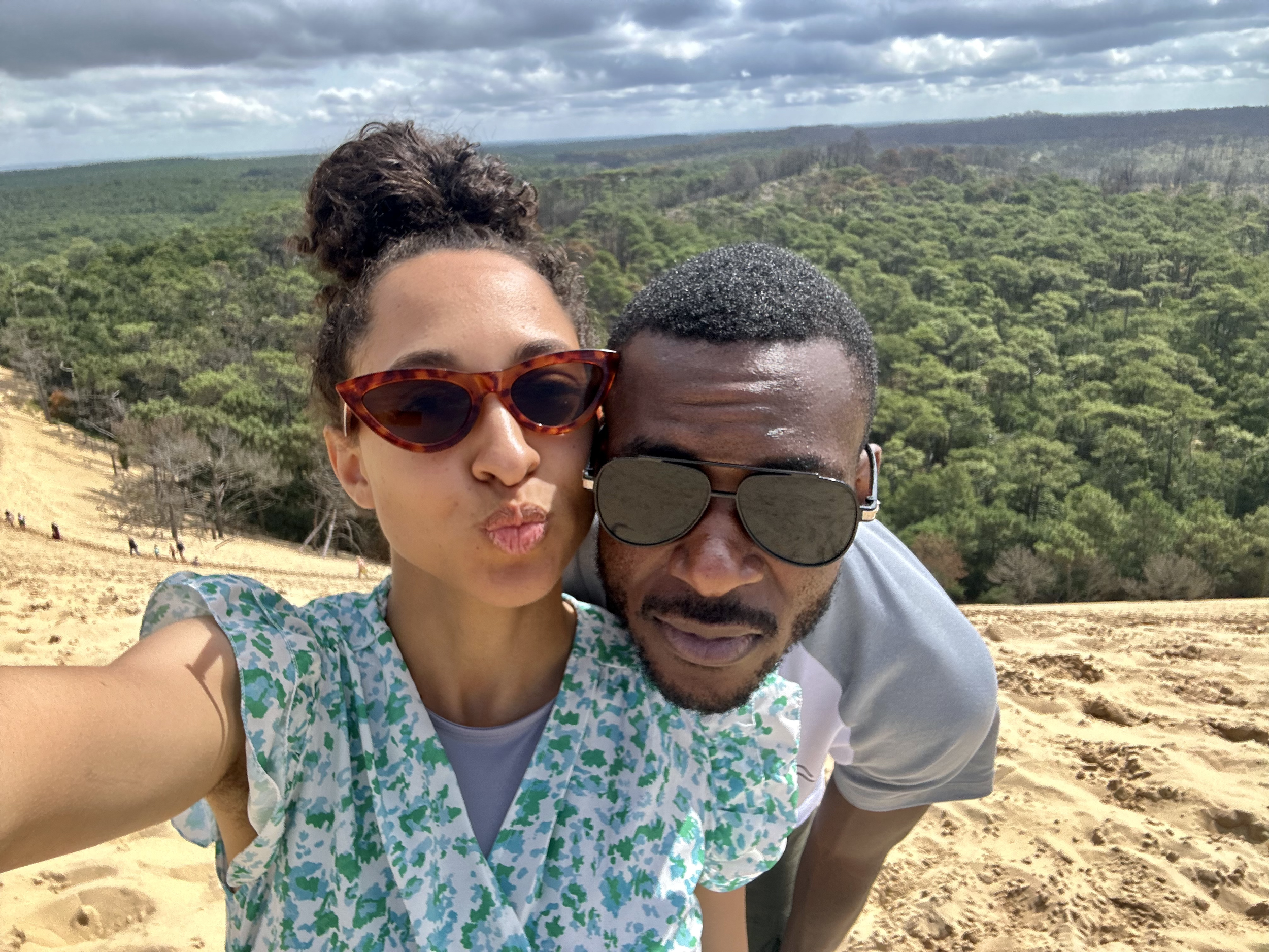 Deux personnes prennent une selfie sur une dune de sable avec une forêt en arrière-plan.