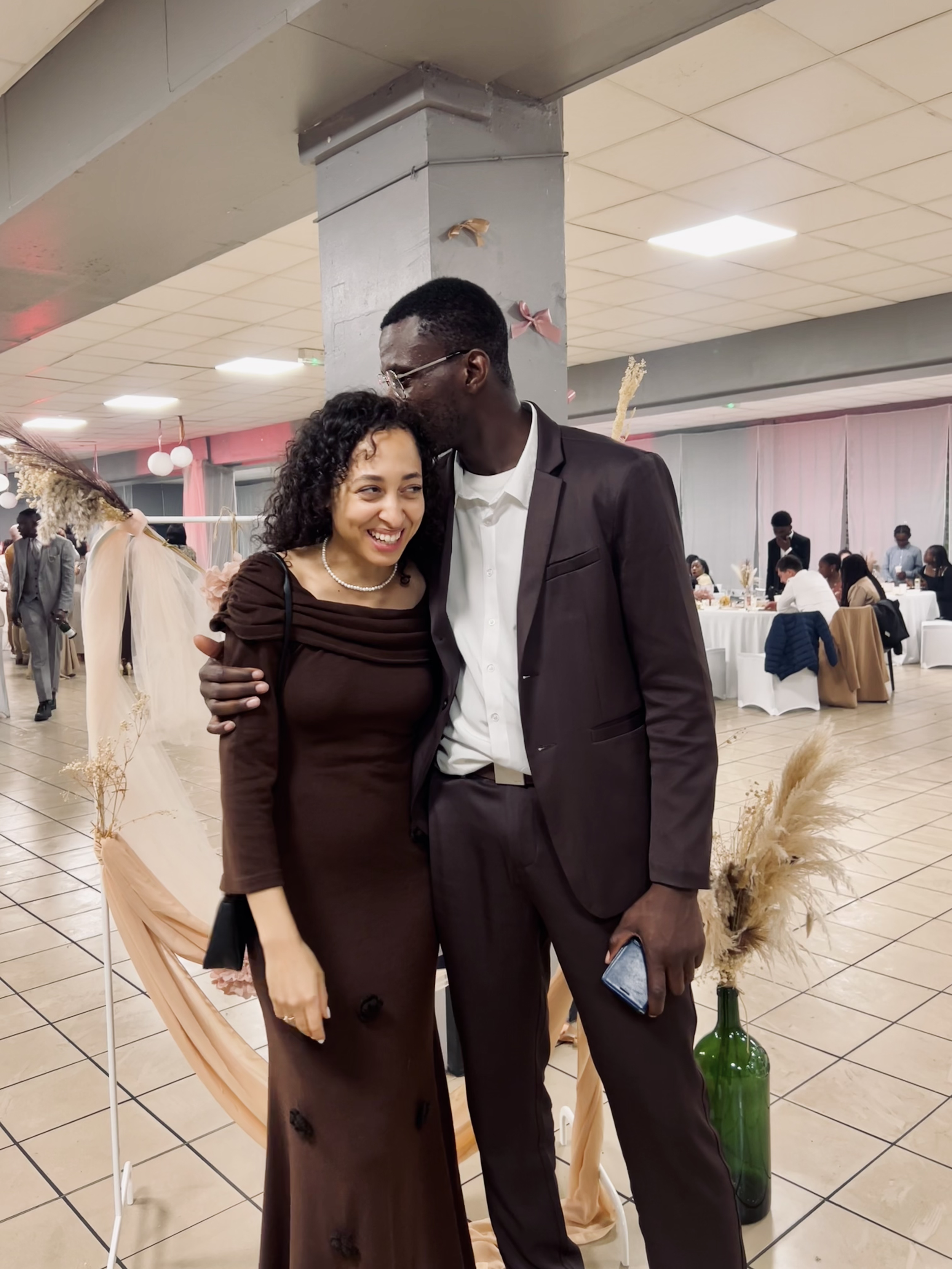 Un couple habillé élégamment se tient dans une salle de réception décorée, entourée de tables et d'invités assis.