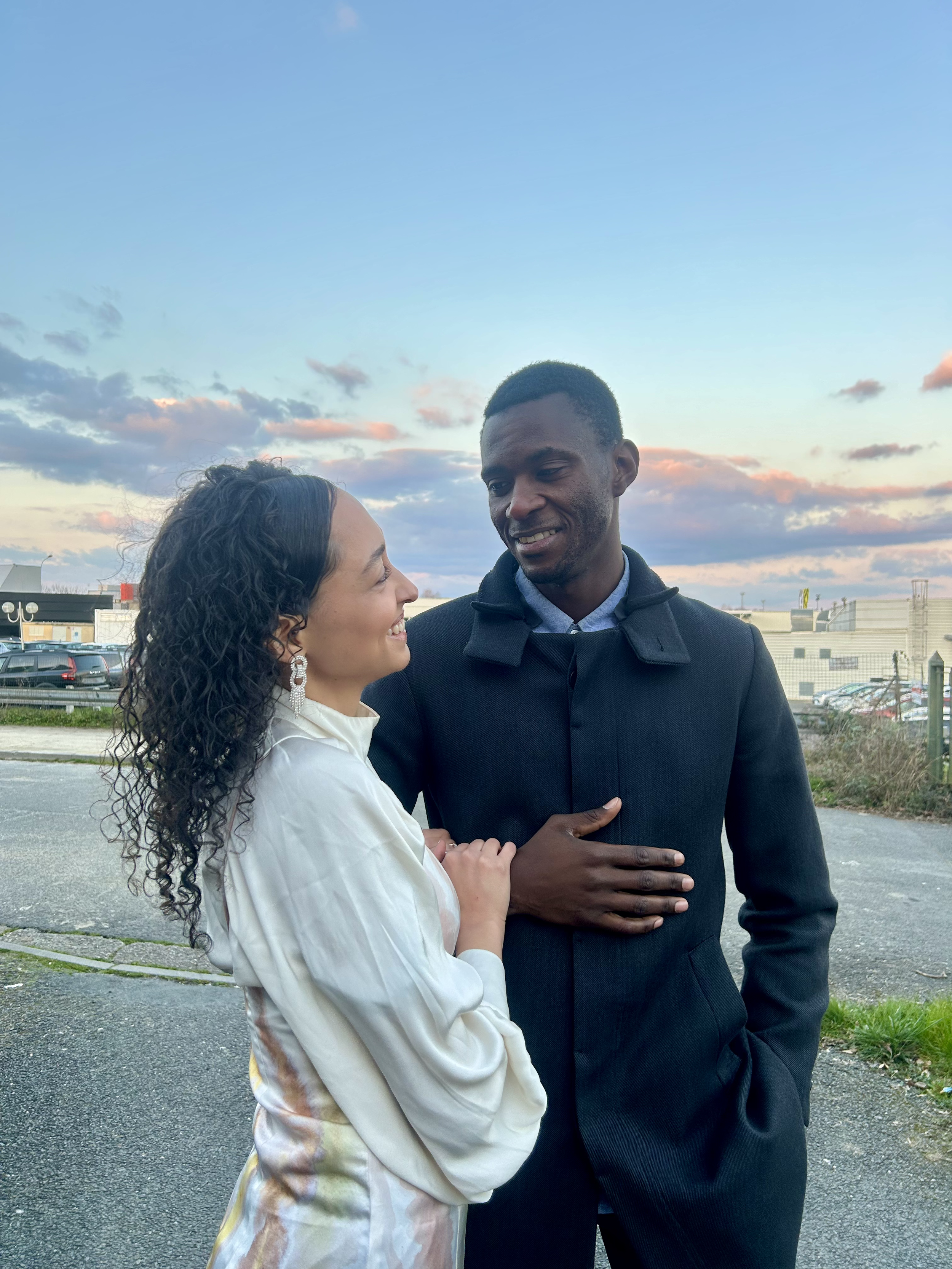 Un homme et une femme souriants l'un à l'autre, debout à l'extérieur avec un ciel coloré en arrière-plan.