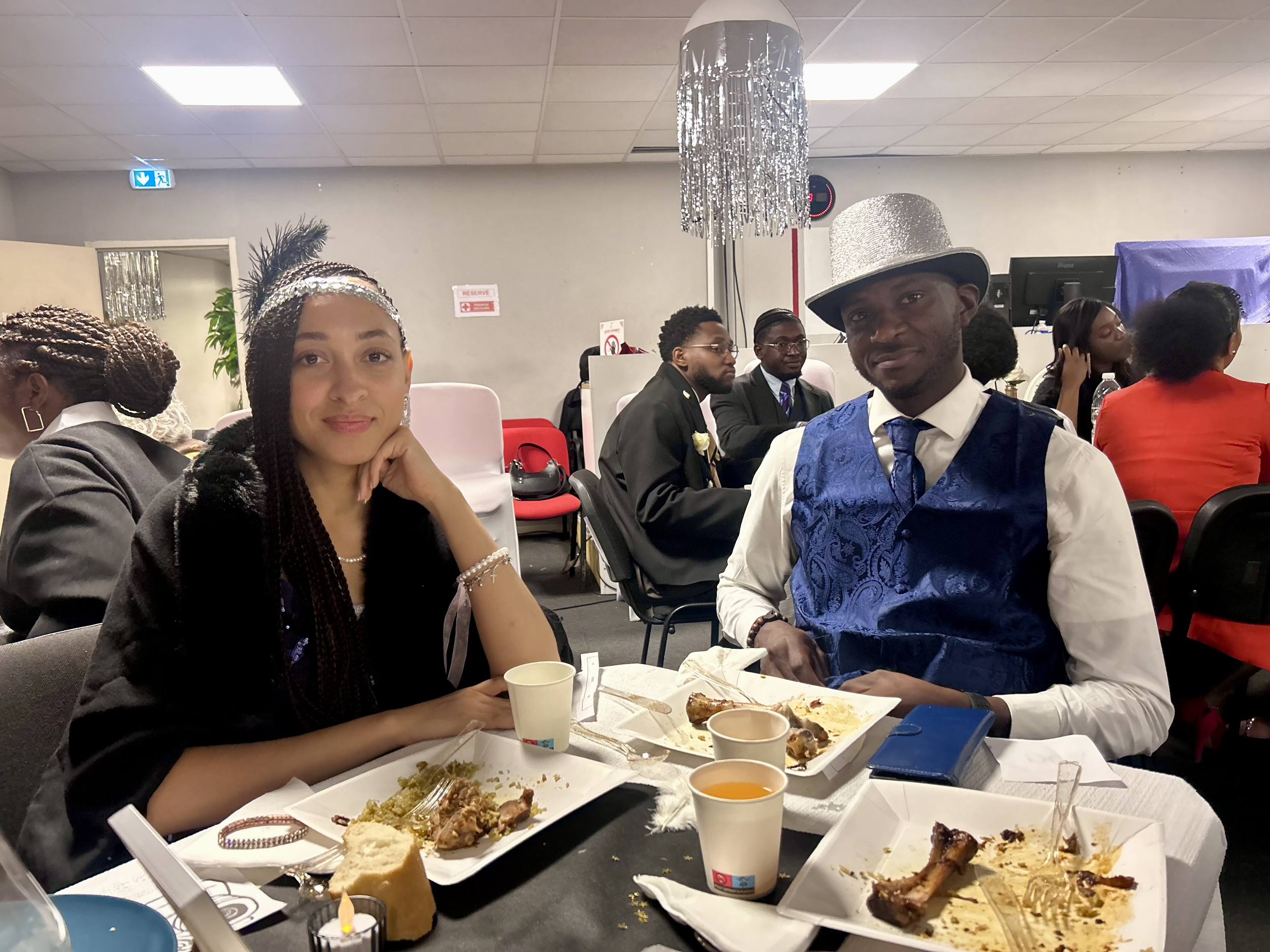 Deux personnes assises à une table lors d'une fête, portant des tenues élégantes avec des accessoires brillants, avec des assiettes vides devant elles.