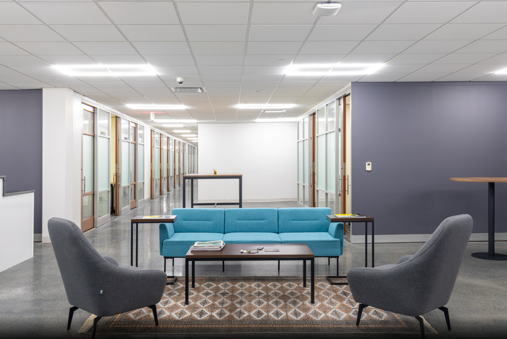Modern office lounge with a turquoise sofa, two gray armchairs, a coffee table on a patterned rug, and hallway with glass doors in the background.