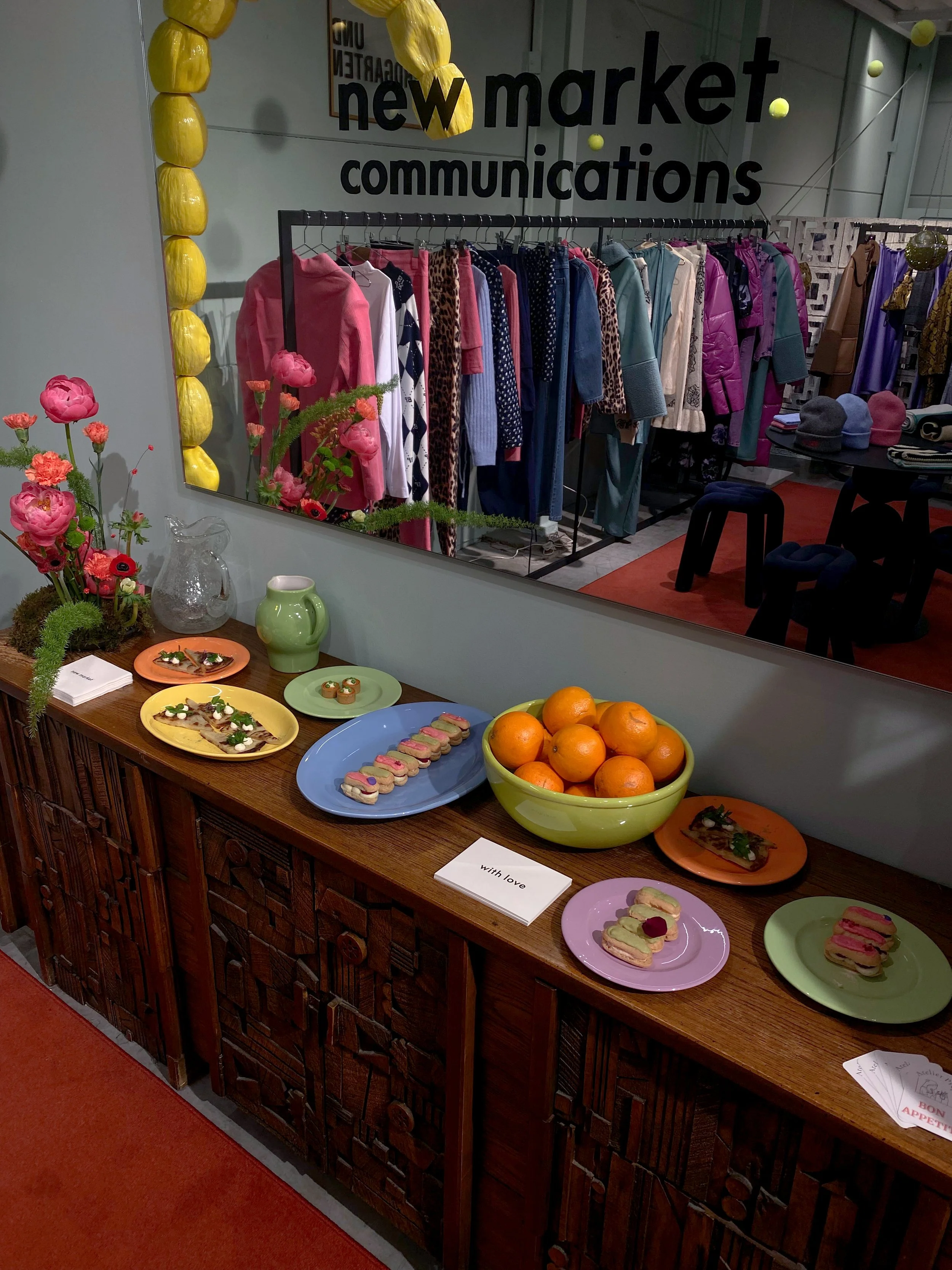 Een tafel met verfijnde hapjes, waaronder sandwiches en kleine taarten, naast een kom met mandarijnen en bloemen in vazen. In de achtergrond zijn kledingrekken met diverse jassen en kleding te zien, evenals een spiegel en een muur met tekst. Een kaartje op de tafel zegt 'with love'.
