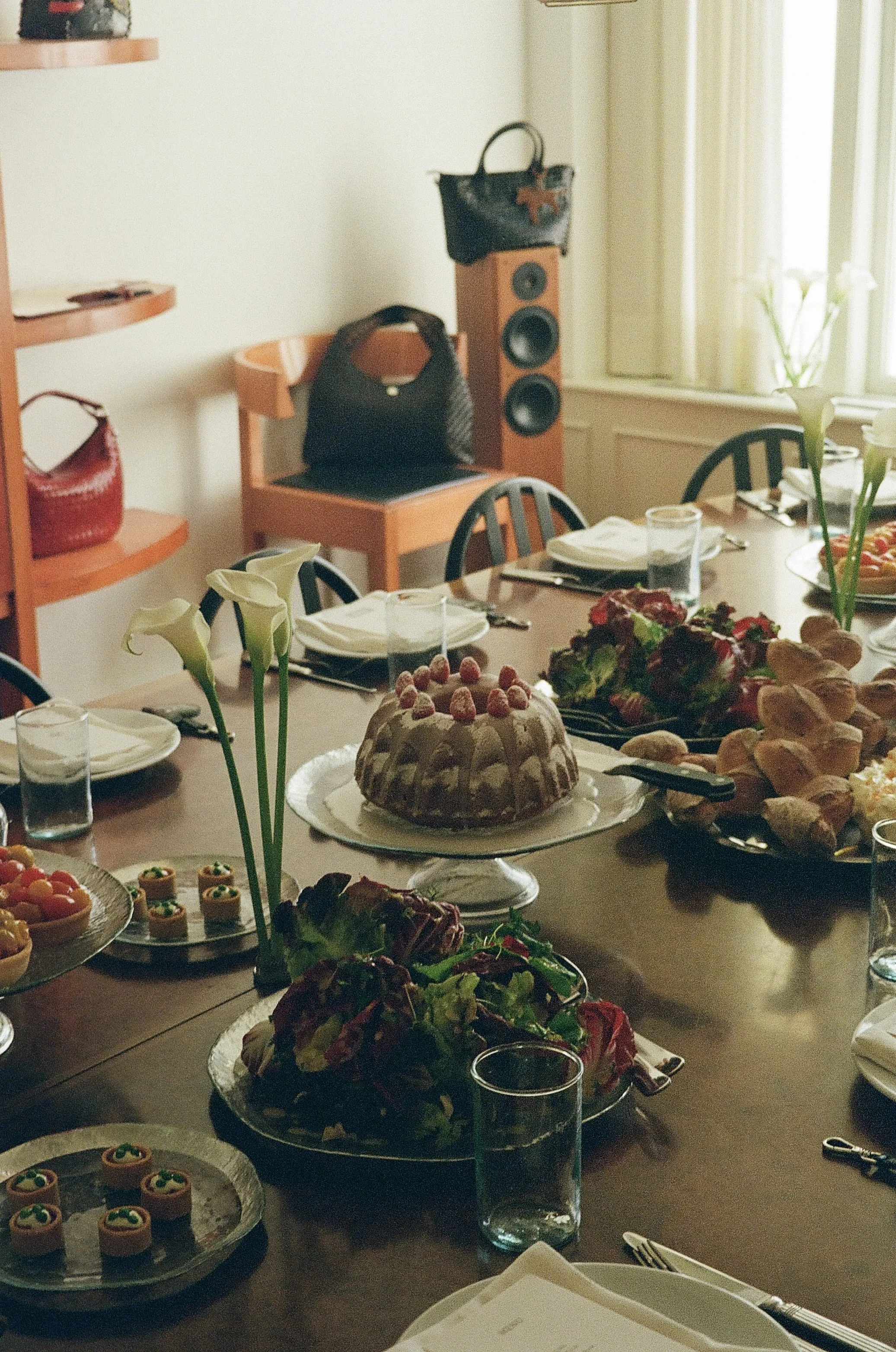 Een feesttafel met verschillende desserts, bloemen en glazen op een donkere houten tafel. In de achtergrond staat een luidspreker en enkele handtassen.