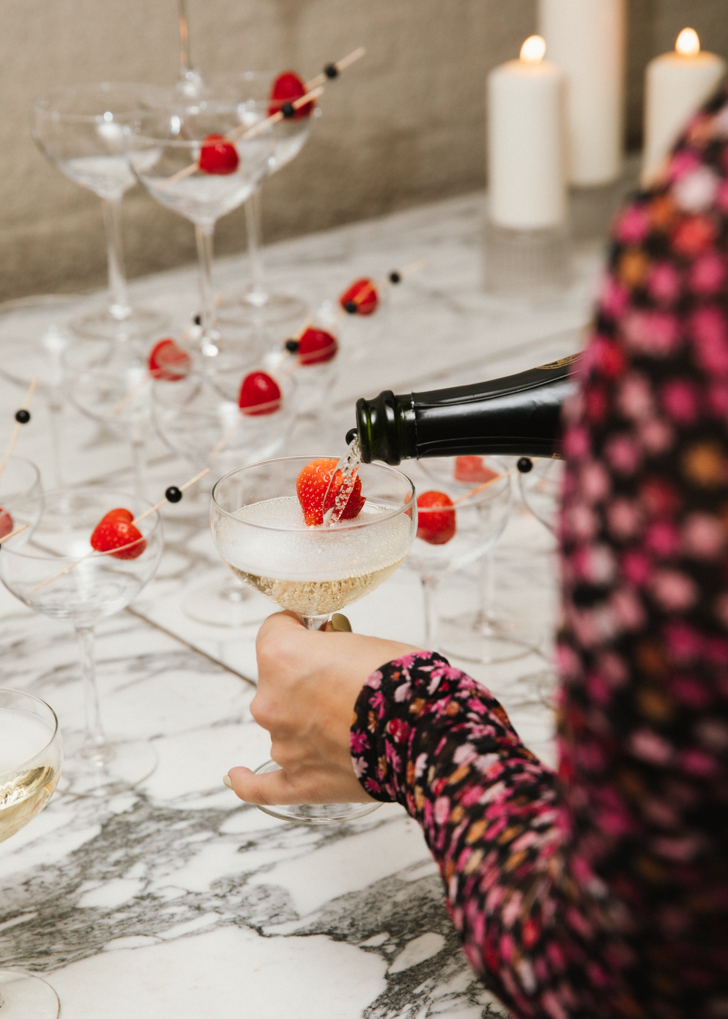 Een persoon met een bloemrijk jasje schenkt champagne in een coupe glas, omringd door champagneglazen met aardbeien en zwarte bessen, met brandende kaarsen op een marmeren tafel.