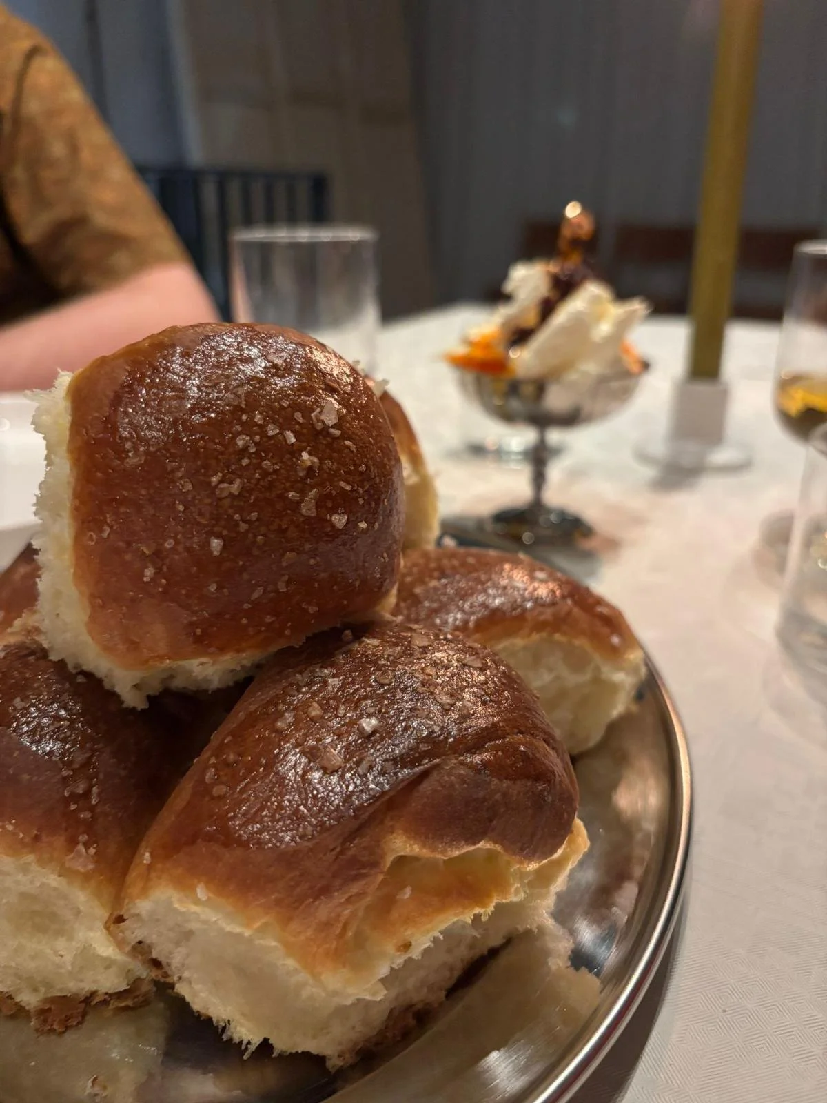 Broodjes op een zilveren schaal met zout, op een tafel met glazen water en een dessert in een coupe met room en chocolade in de achtergrond.