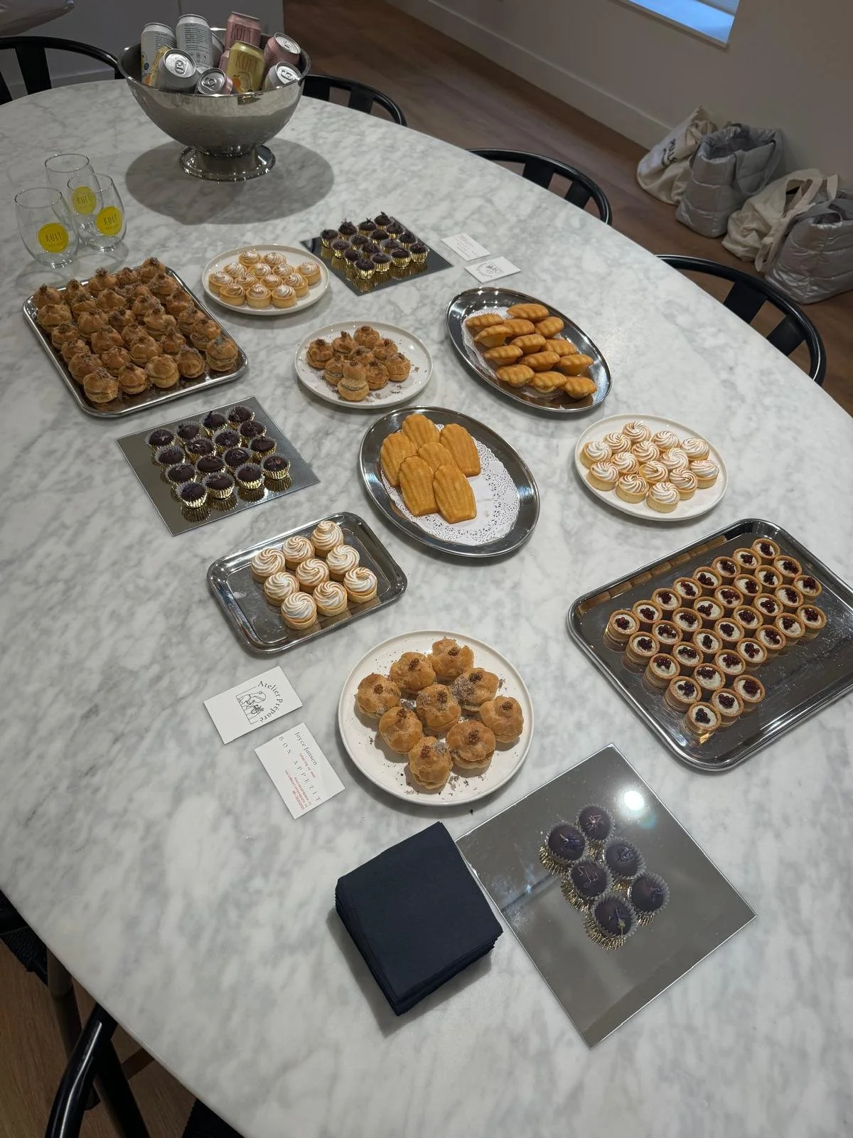 Een ronde marmeren tafel met diverse kleine gebakjes en koekjes, waaronder cupcakes,ètrets, en mini-tartlets, en een grote kom met blikjes drinken, in een lichte kamer met houten vloer en ramen.